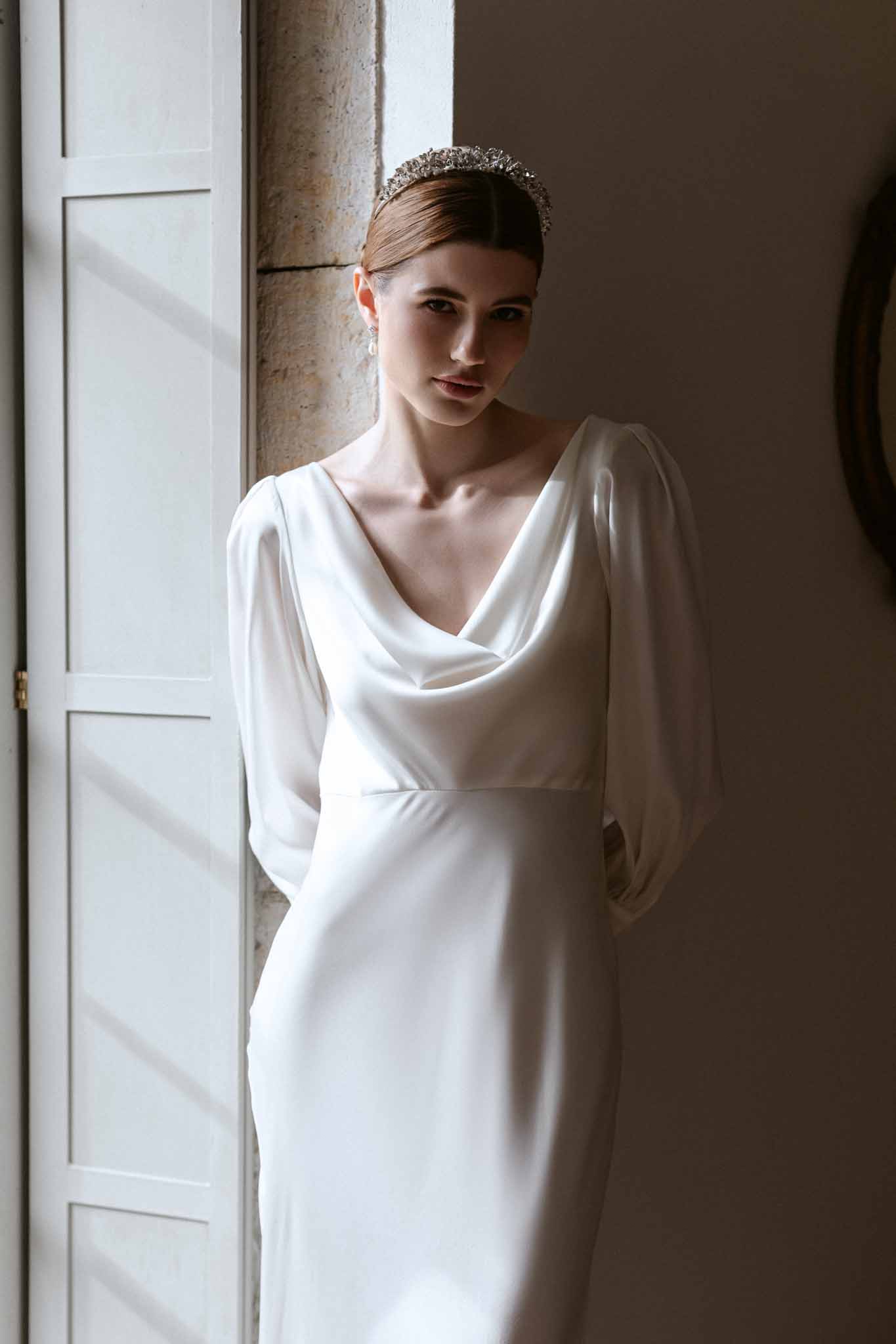 Bride in cowl-neck ivory gown with crystal tiara and drop earrings standing beside a white paneled door