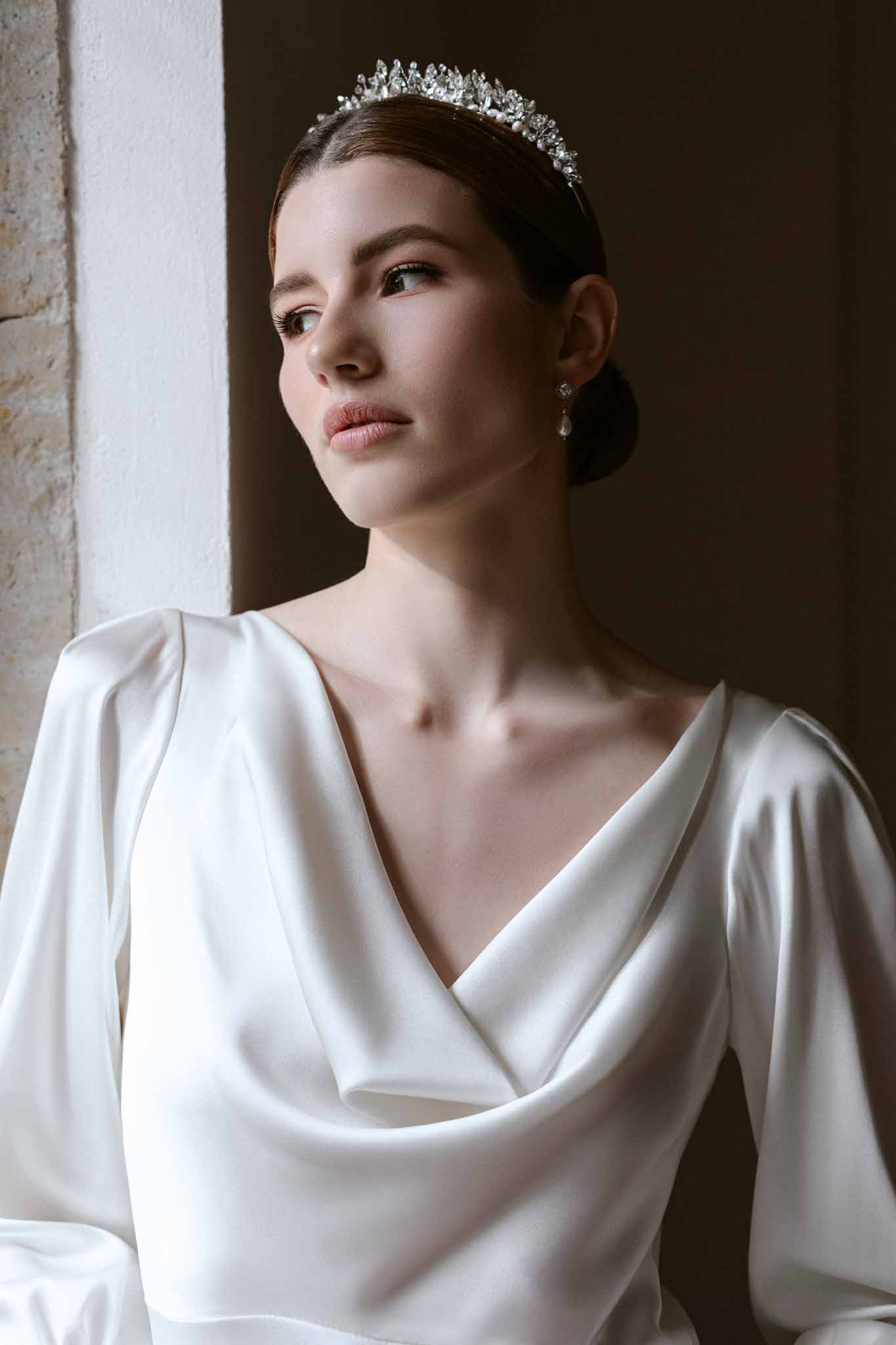 Bride in profile wearing minimalist ivory silk cowl-neck gown with crystal tiara and pearl earrings against dark backdrop