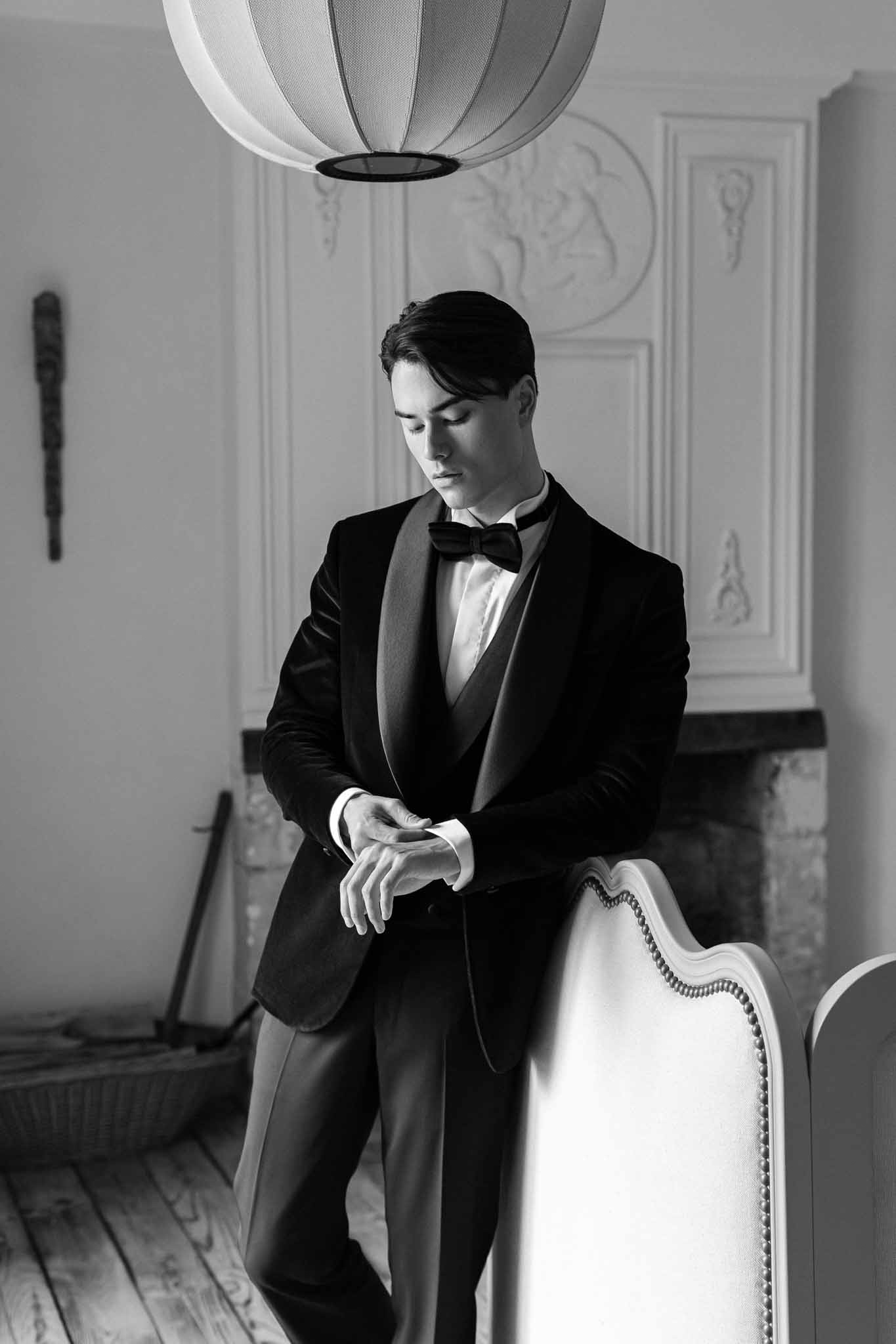 Black-and-white portrait of groom in black tuxedo with white gloves seated in ornate panelled interior