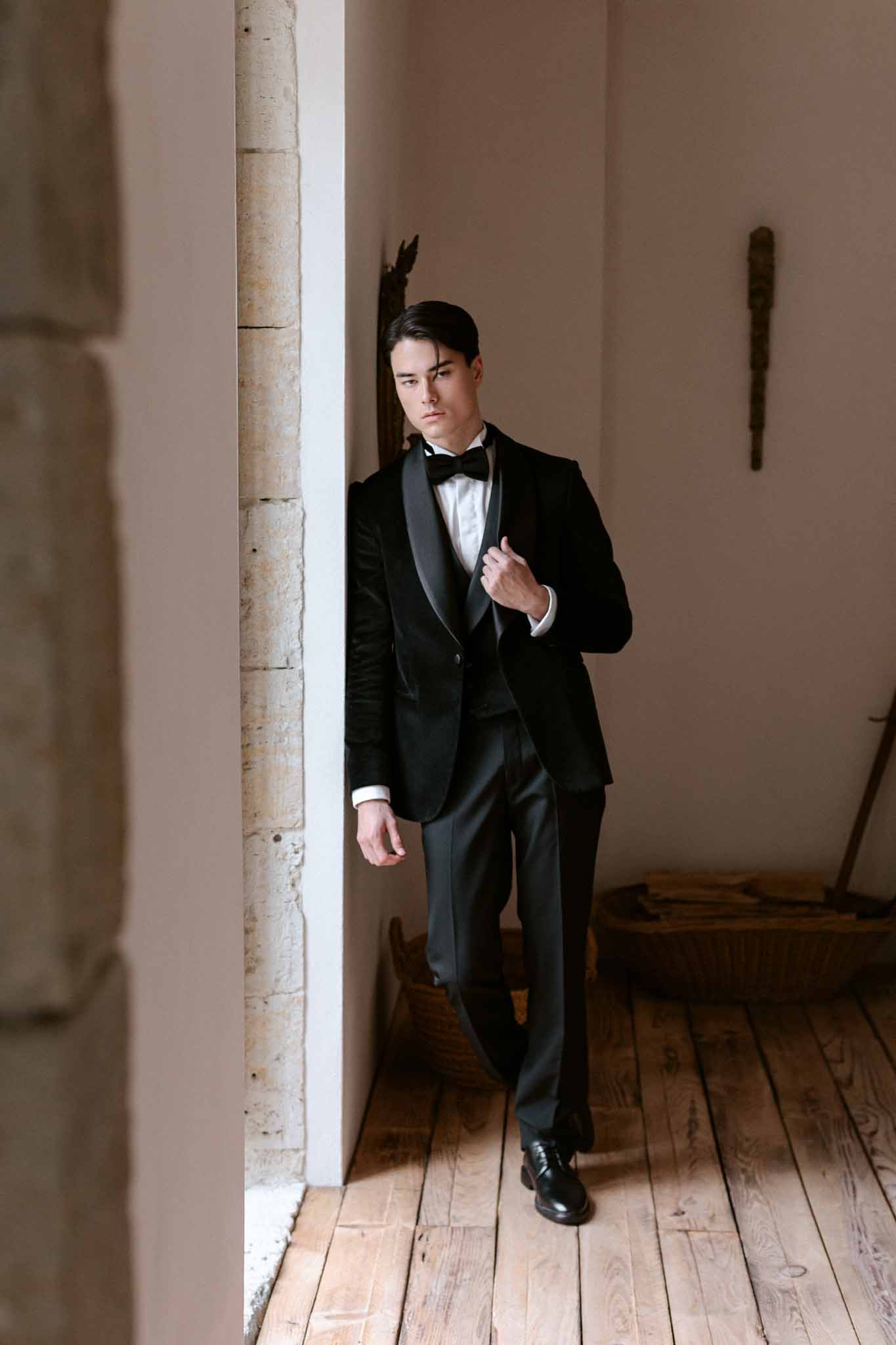 Groom in black tuxedo with bow tie posing in full-length portrait against cream walls and honey-toned floor in corridor