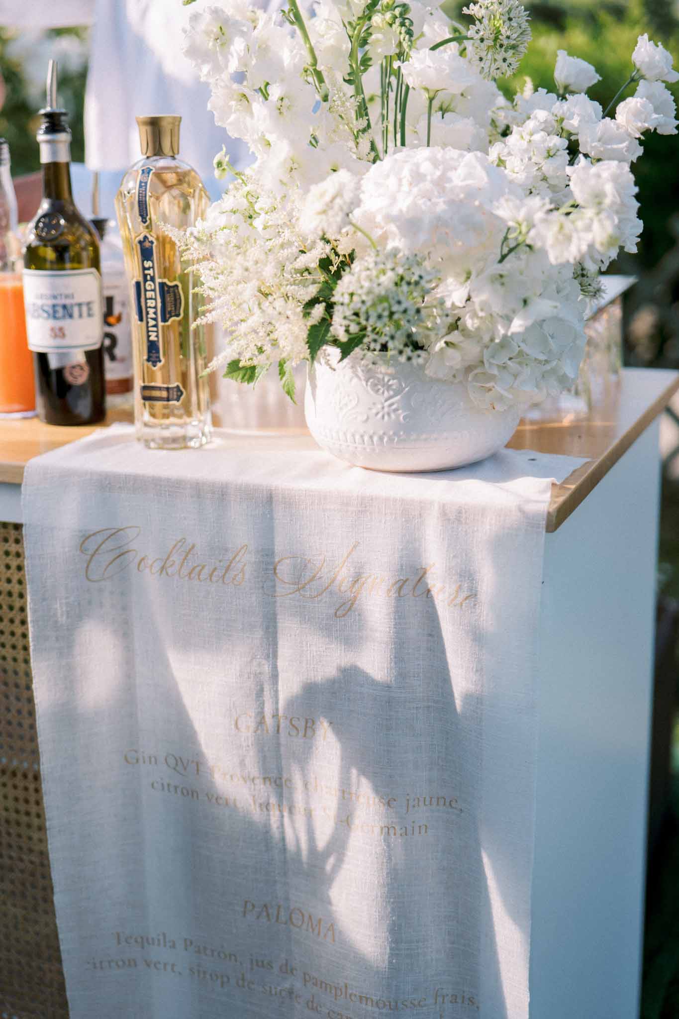 Cocktail bar with gold calligraphy menu listing signature drinks, white hydrangea arrangement, and spirit bottles