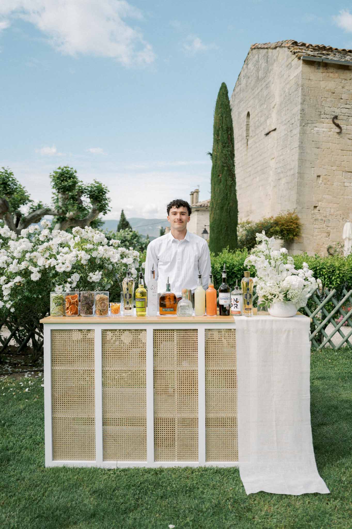 Bartender behind a white cane-panel cocktail bar on a lawn with hydrangeas and a Provencal stone building behind