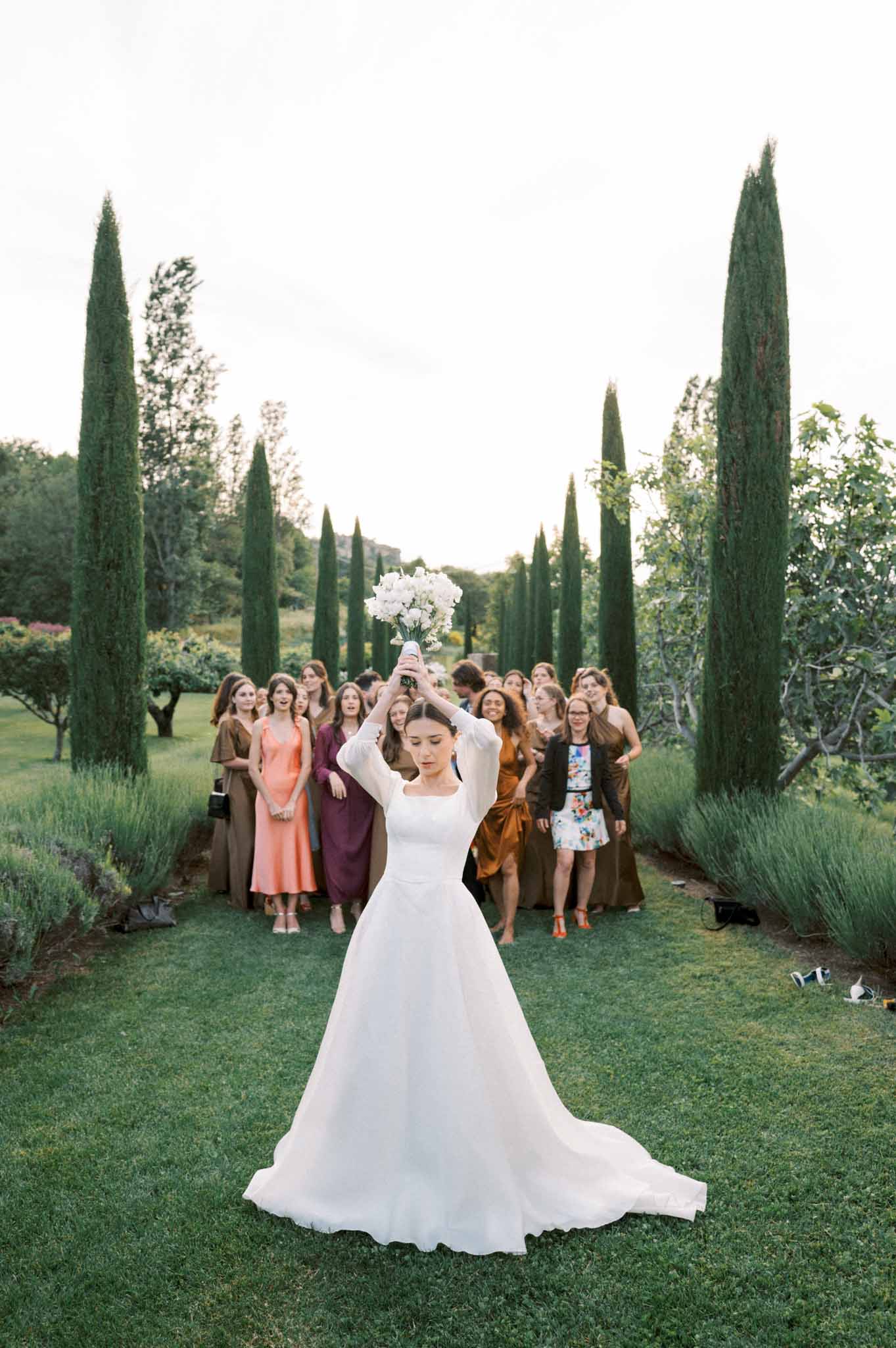 Bride tossing white bouquet in cypress and lavender allee with fifteen female guests gathered behind