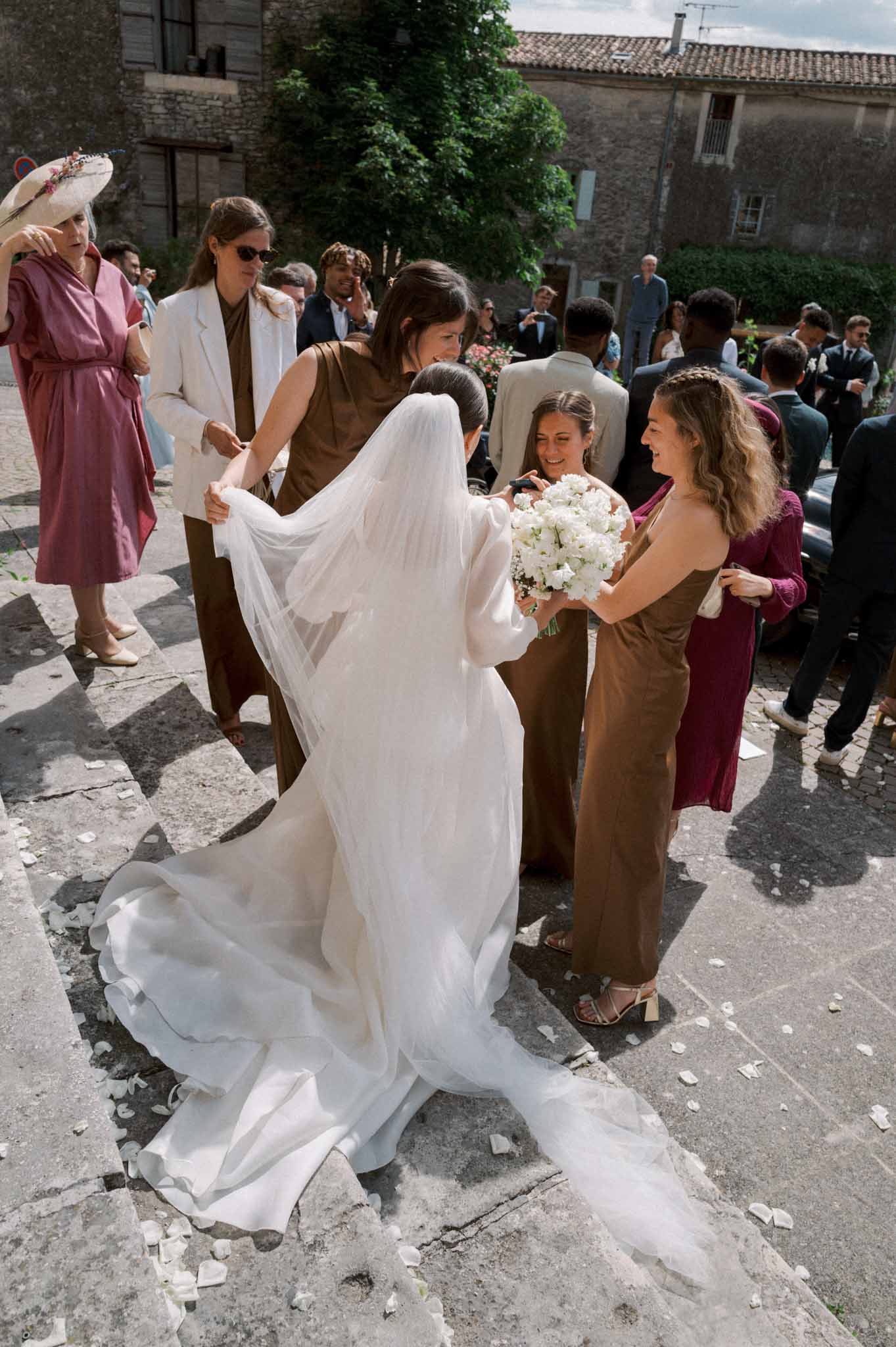 Bride from behind with cathedral veil as bridesmaids in olive dresses arrange veil amid scattered petals