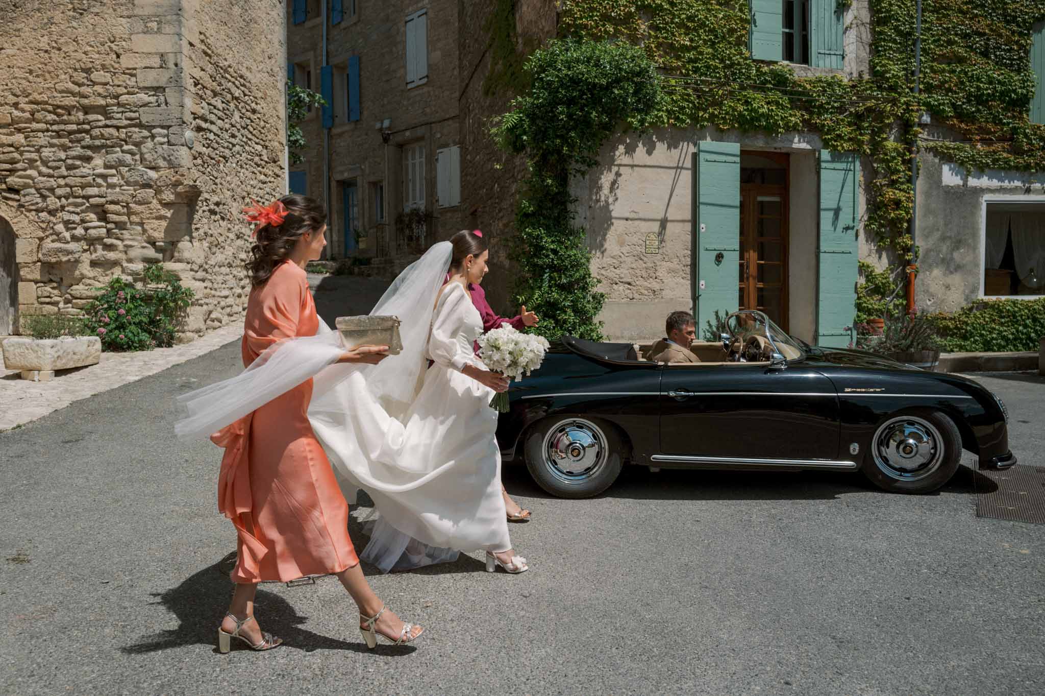 Bride with cathedral veil and two attendants in coral dresses beside vintage black Porsche in Provencal village