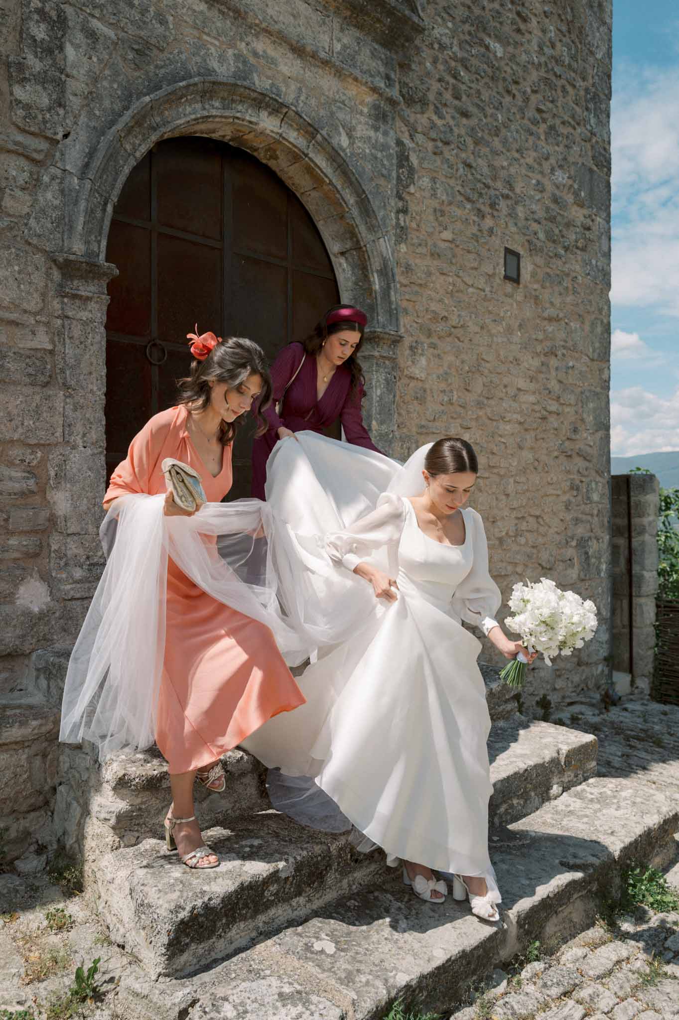 Bride in white satin gown descending stone chapel steps assisted by two guests managing her train and veil