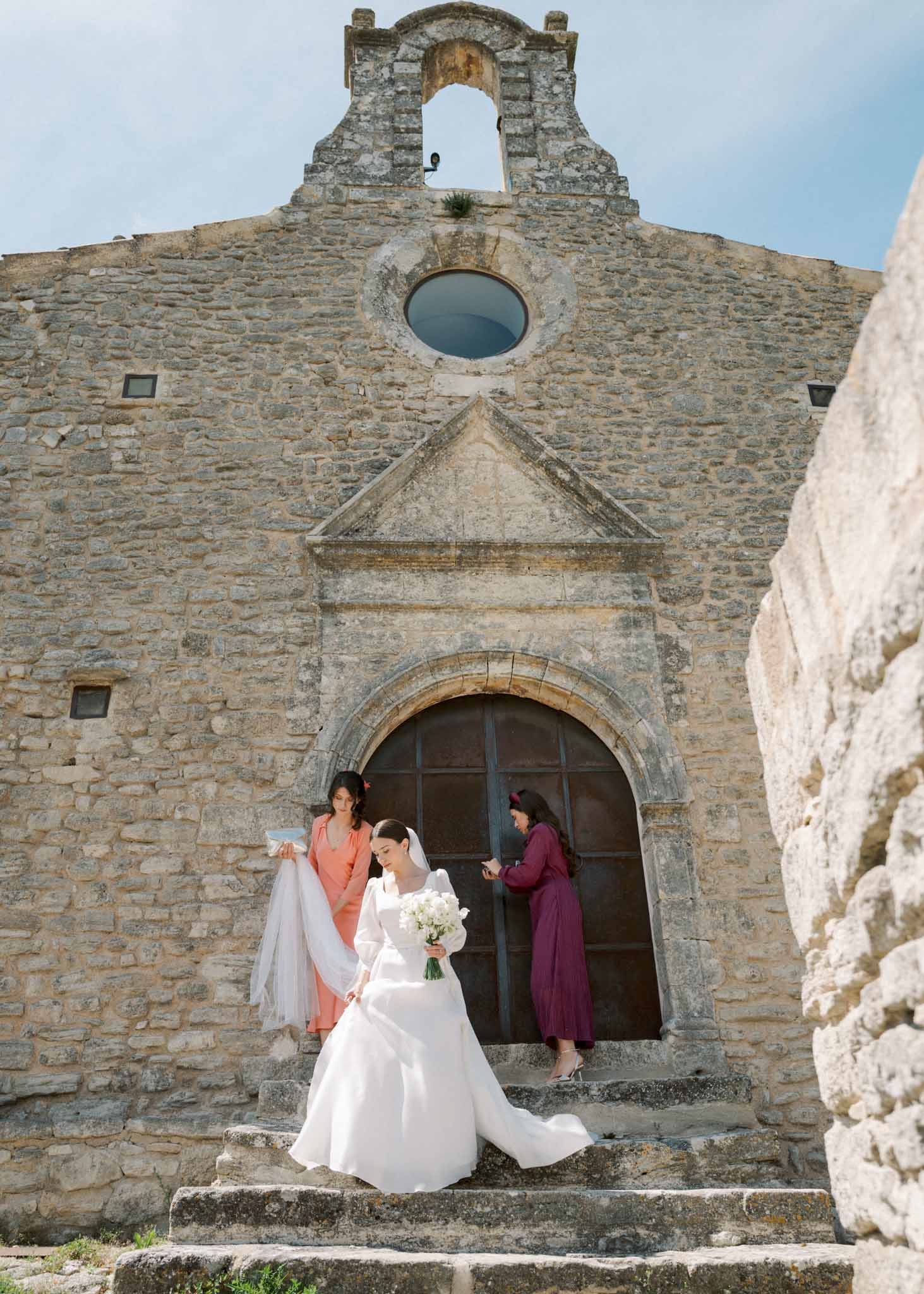 Bride with cathedral veil and white bouquet on Romanesque chapel steps as two attendants assist