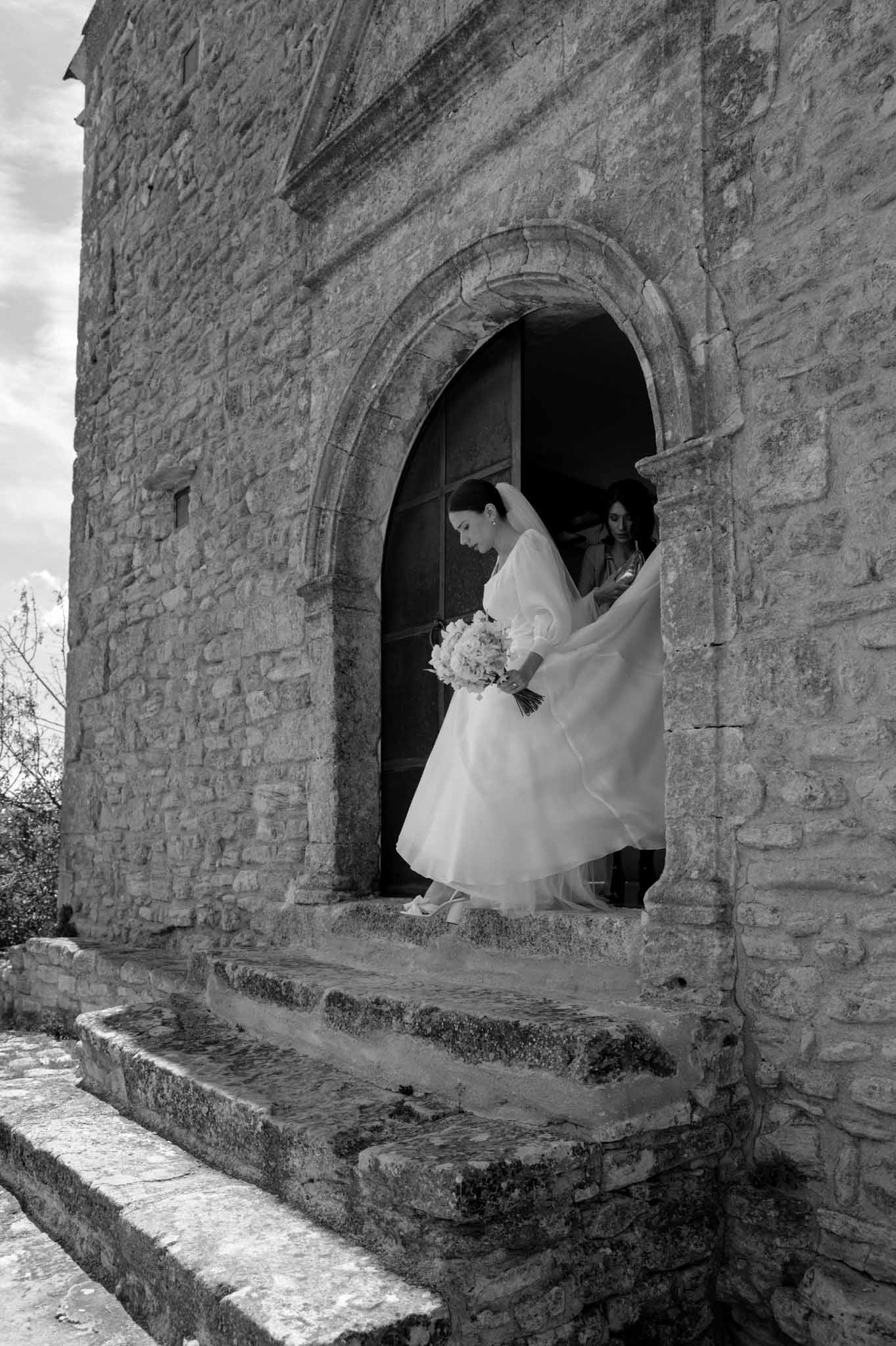 Bride exits Romanesque chapel arch with wind-caught veil and rounded bouquet as attendant assists in B&W