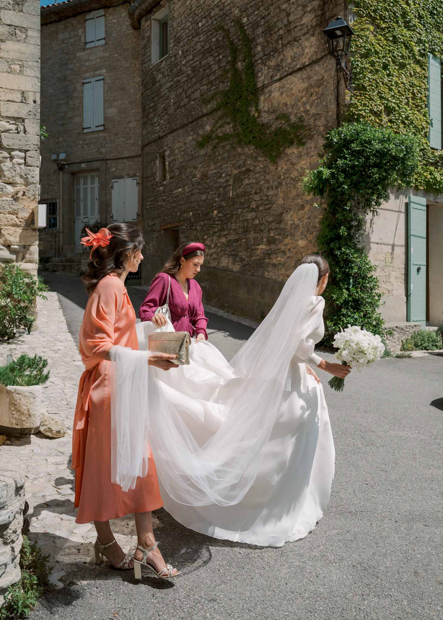 Bride in ivory gown with cathedral veil carried by two guests walking through Provencal village street