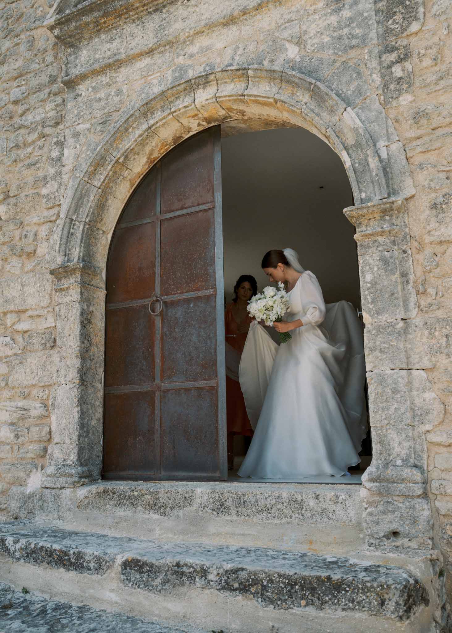 Bride stepping through Romanesque stone archway in puff-sleeve gown with white peony bouquet