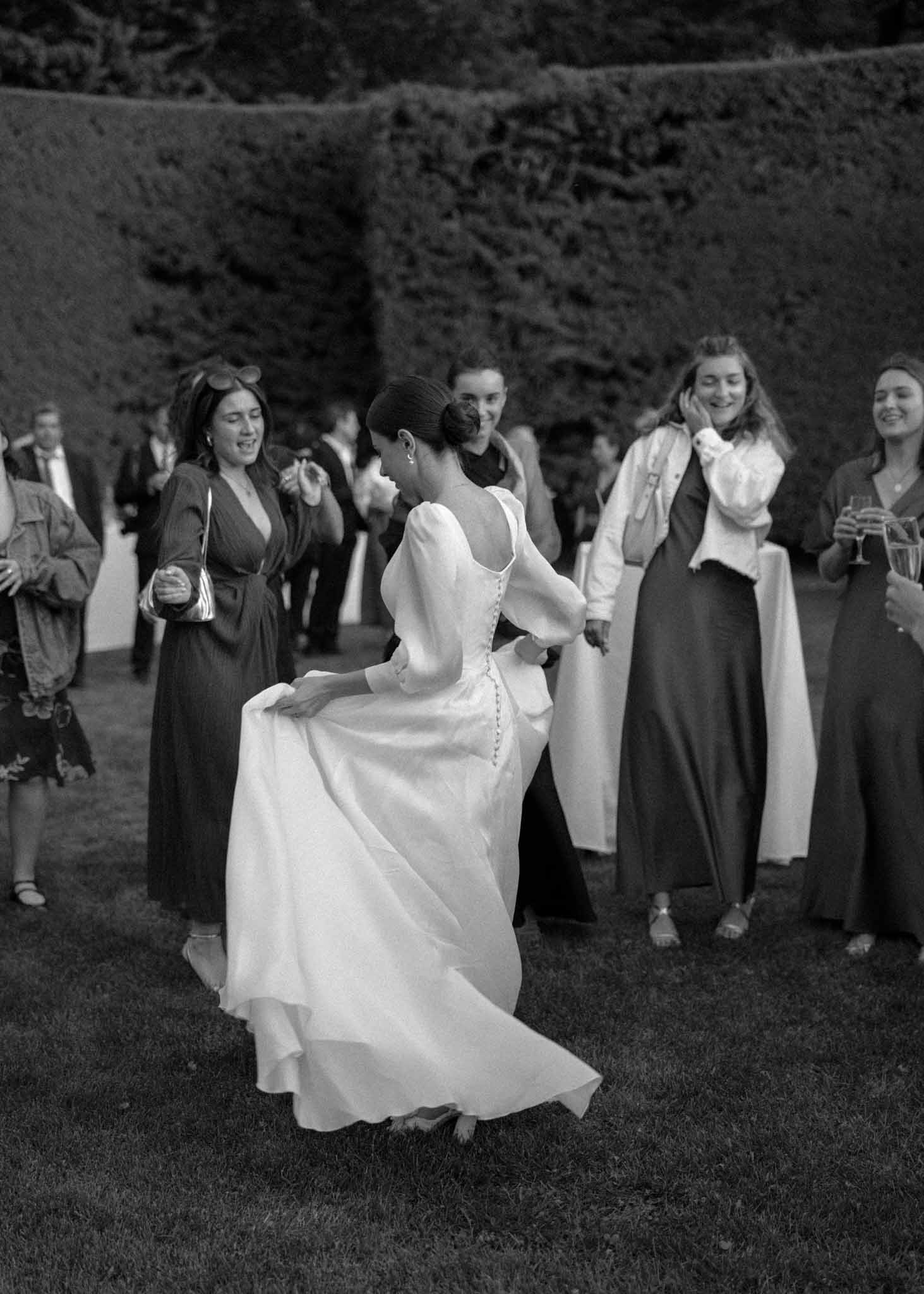 Black-and-white shot of bride dancing barefoot on lawn surrounded by laughing guests near clipped hedges