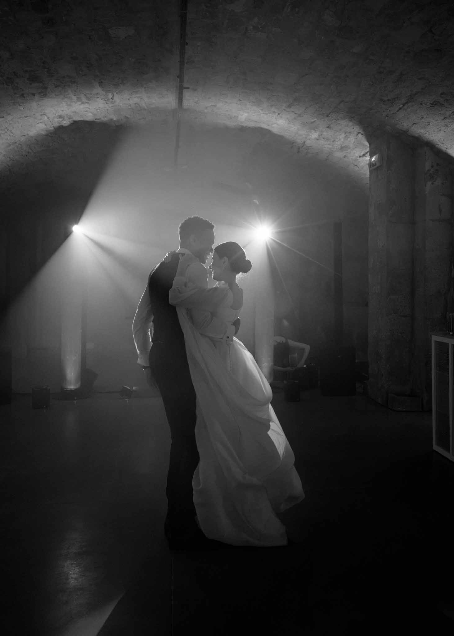 Black and white first dance with spotlight beams through haze in vaulted stone cellar