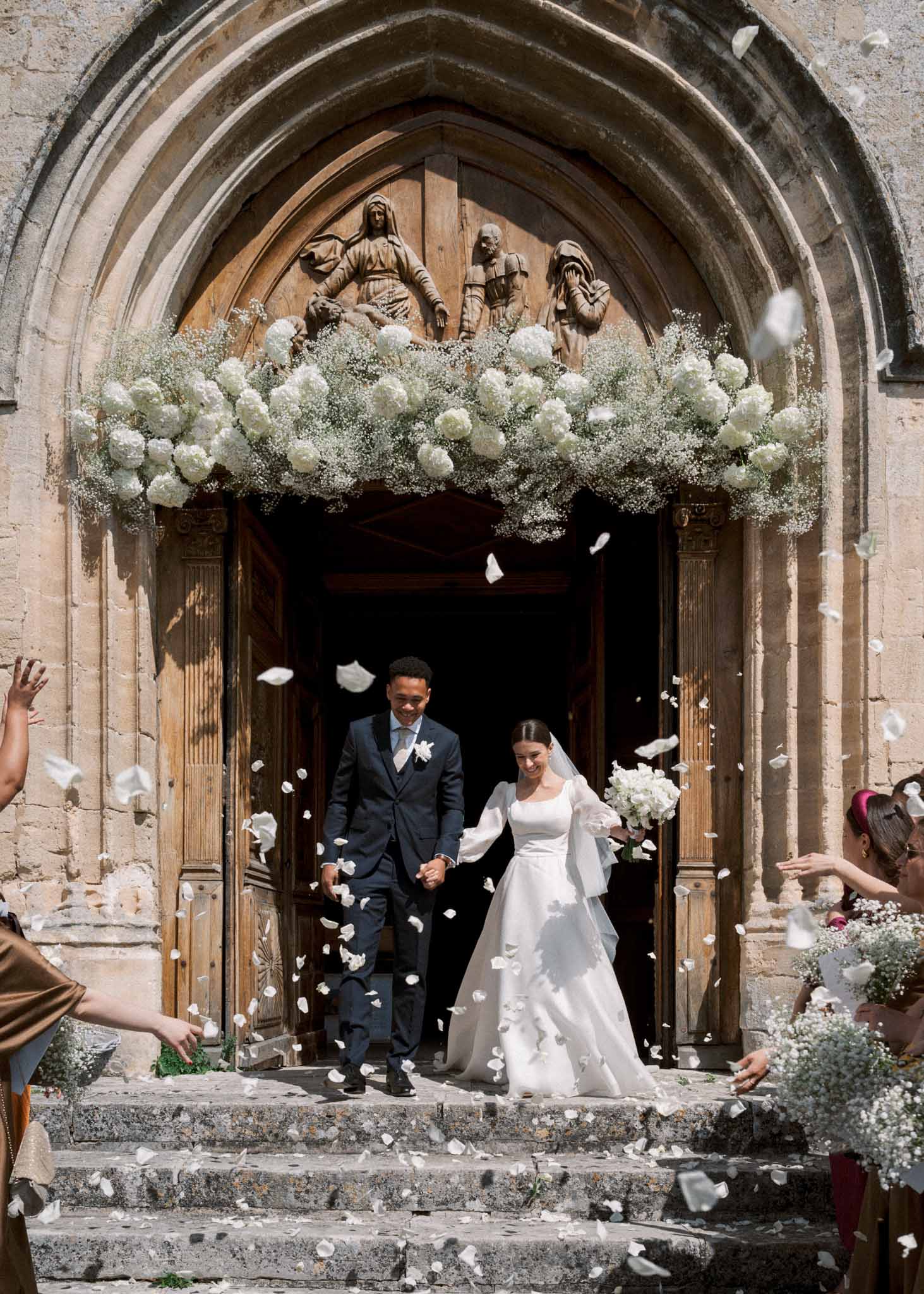 Bride and groom exit a Gothic stone chapel as guests throw white petals under a hydrangea garland arch