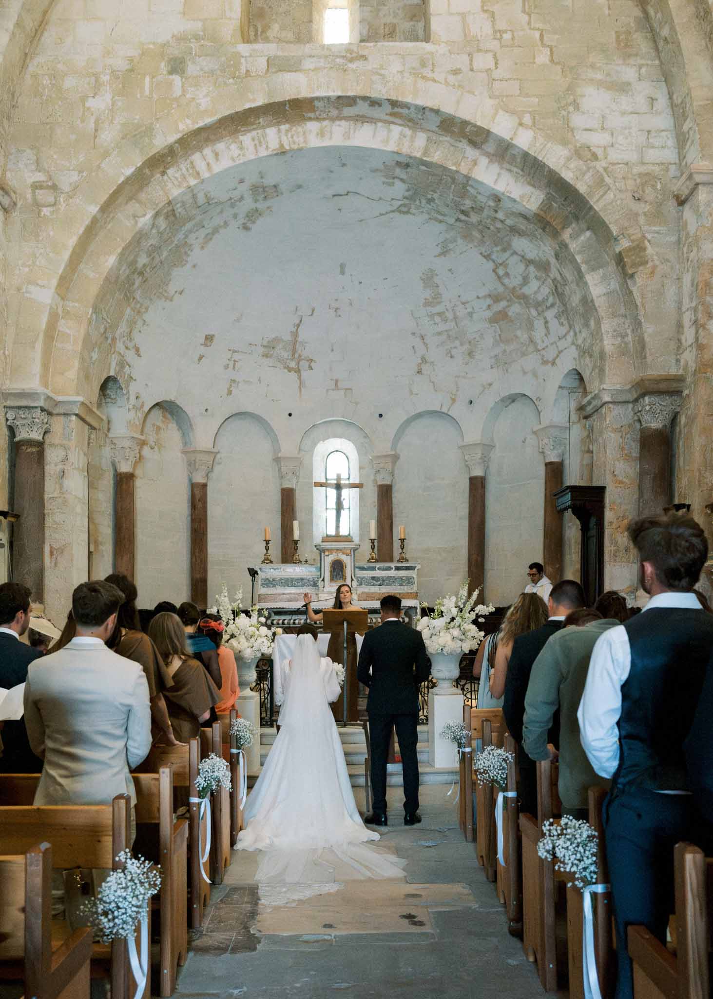 Wedding ceremony inside Romanesque stone chapel with bride and groom at altar flanked by white flower arrangements