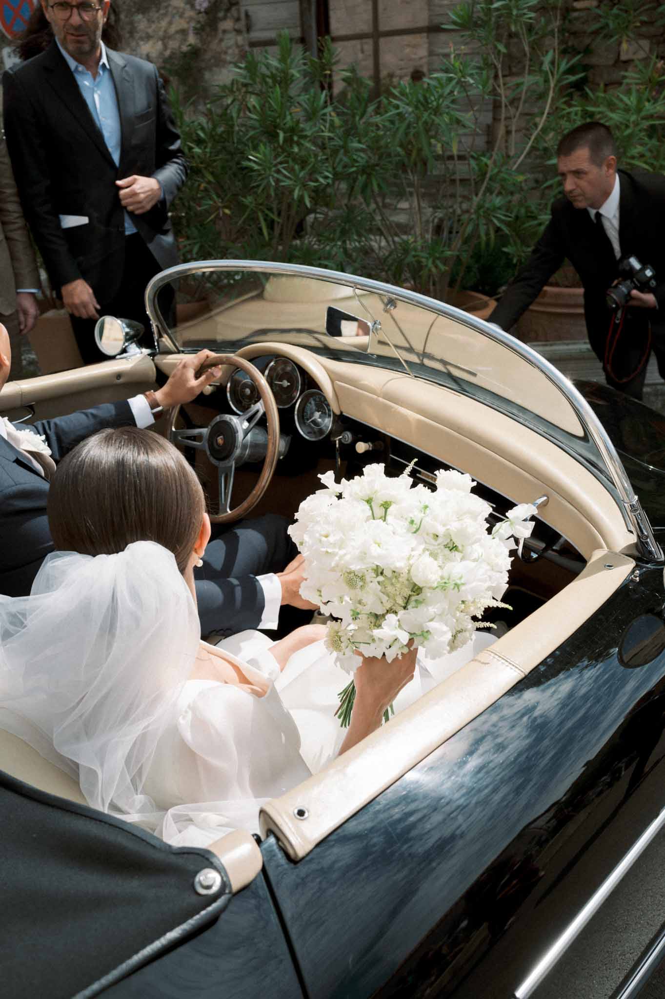 Overhead shot of couple in vintage green Porsche convertible, bride holding all-white peony bouquet