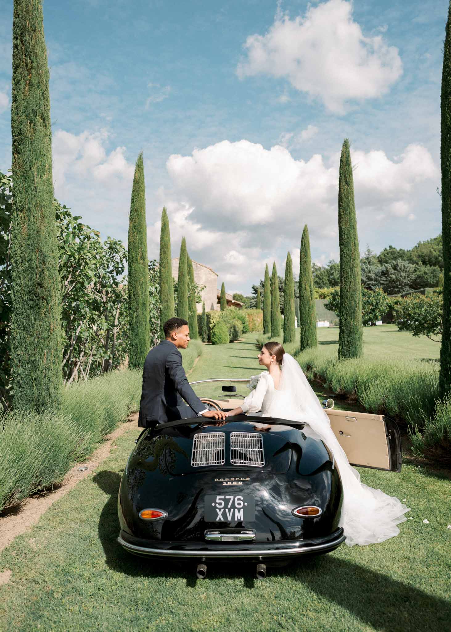 Bride and groom seated in vintage black Porsche convertible on cypress-lined driveway with lavender borders
