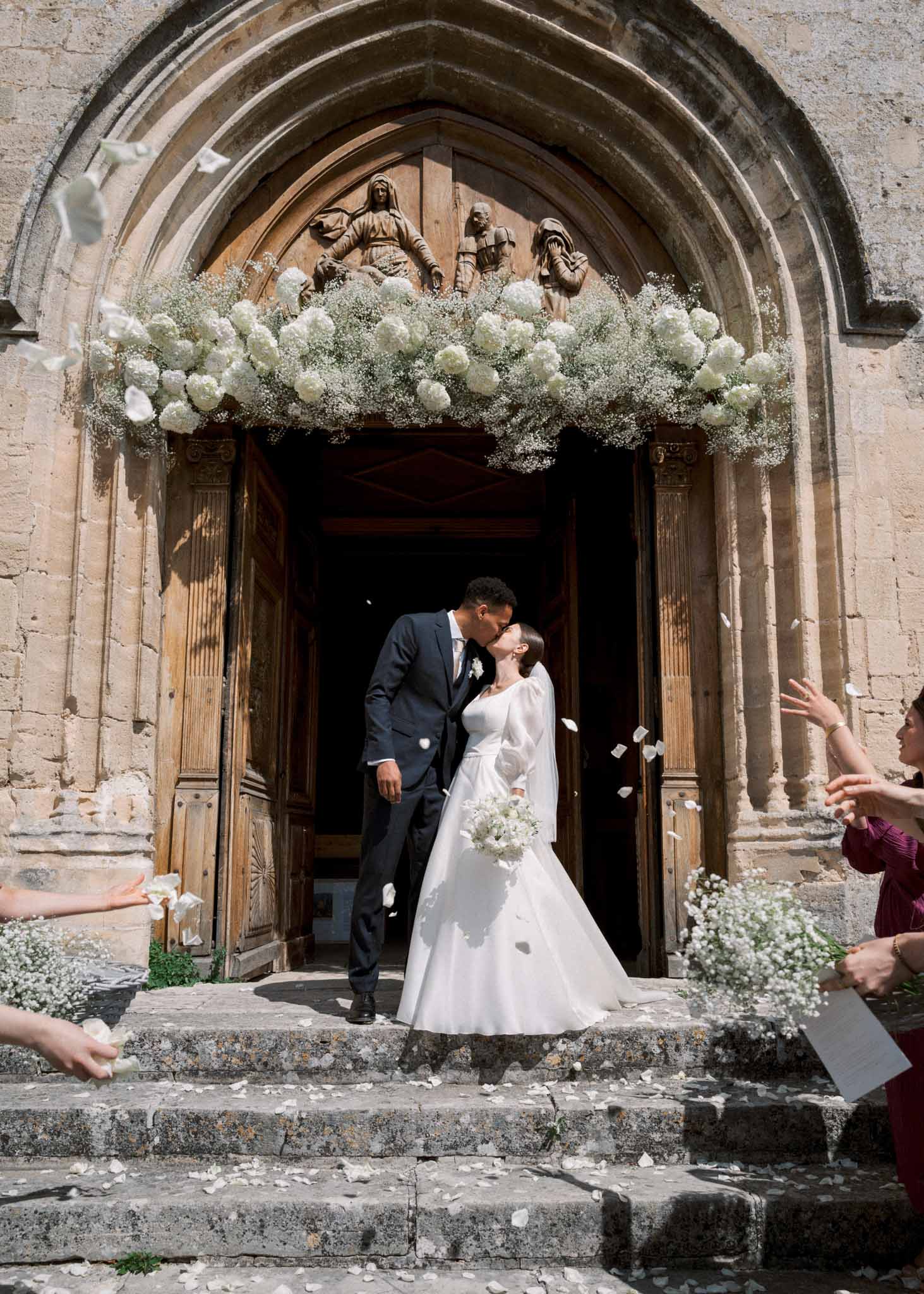 Bride and groom kiss under Romanesque chapel doorway adorned with white hydrangeas as guests toss flower petals