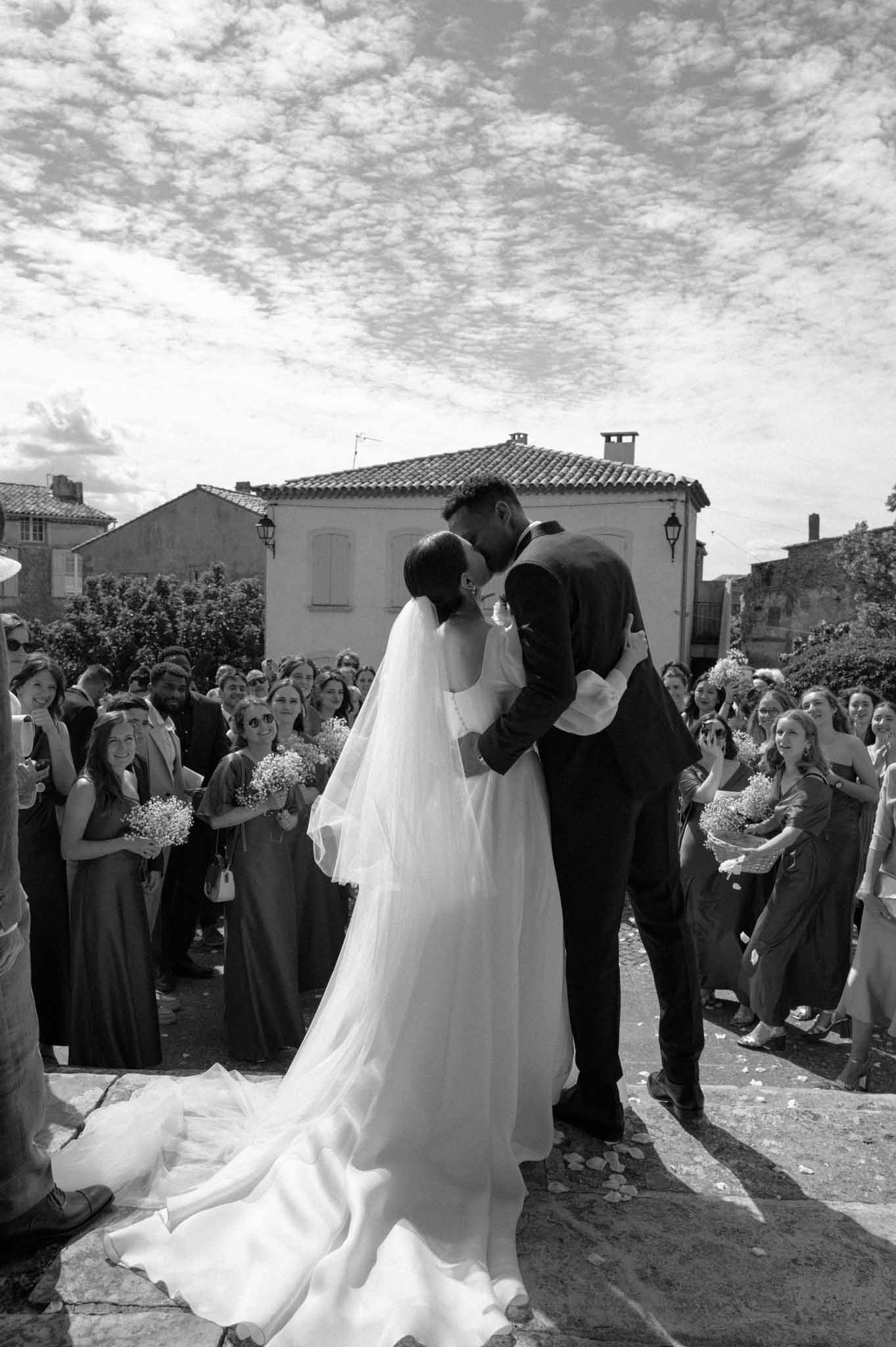 Bride and groom first kiss on church steps with scattered petals and guests lining both sides in B&W