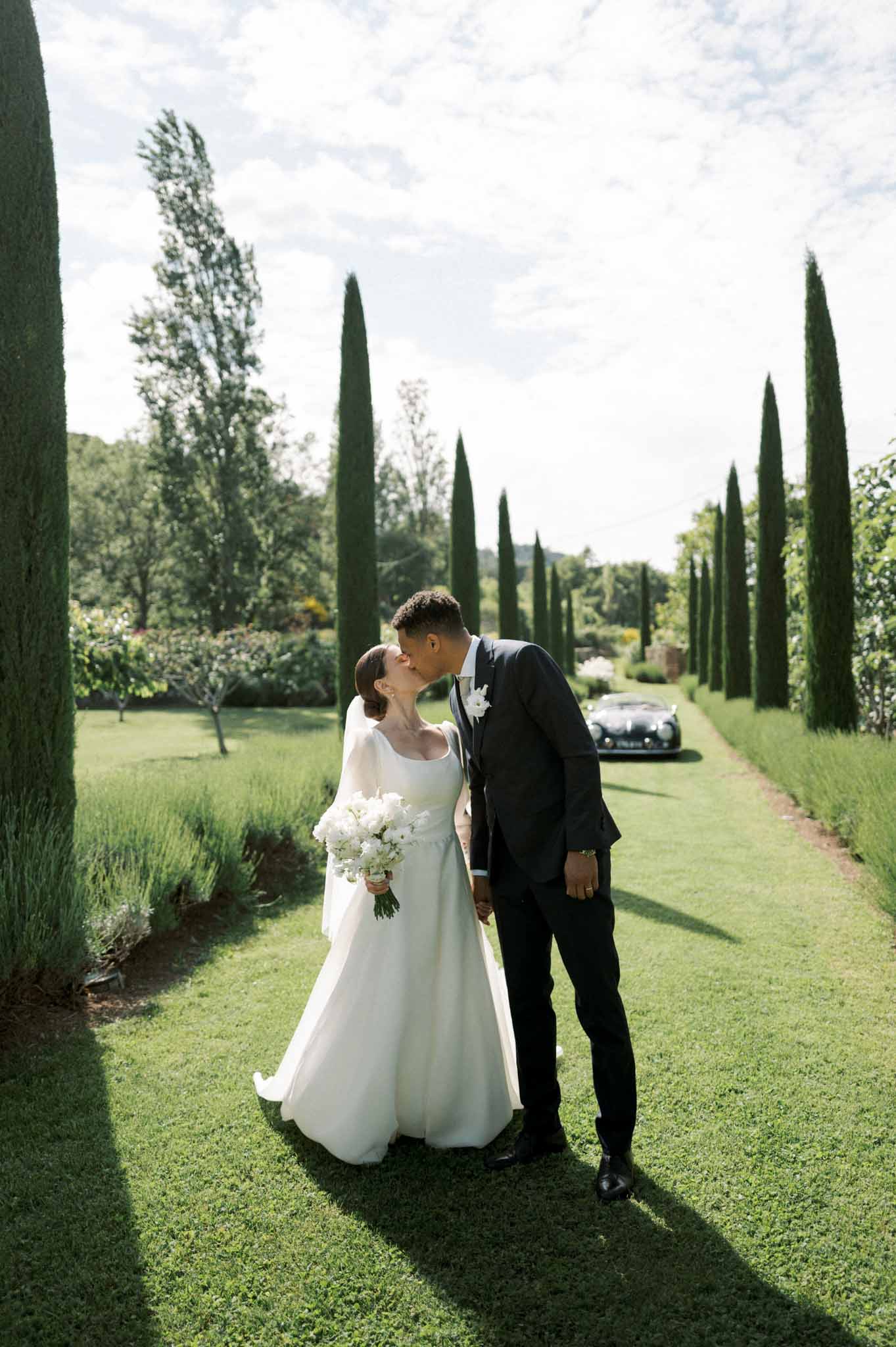 Bride and groom kissing on cypress-lined lawn allee with vintage sports car in background