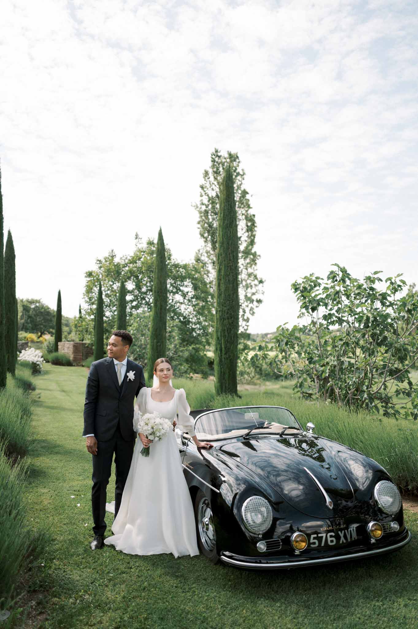 Bride and groom standing beside a vintage black Porsche convertible in a formal garden with cypress trees