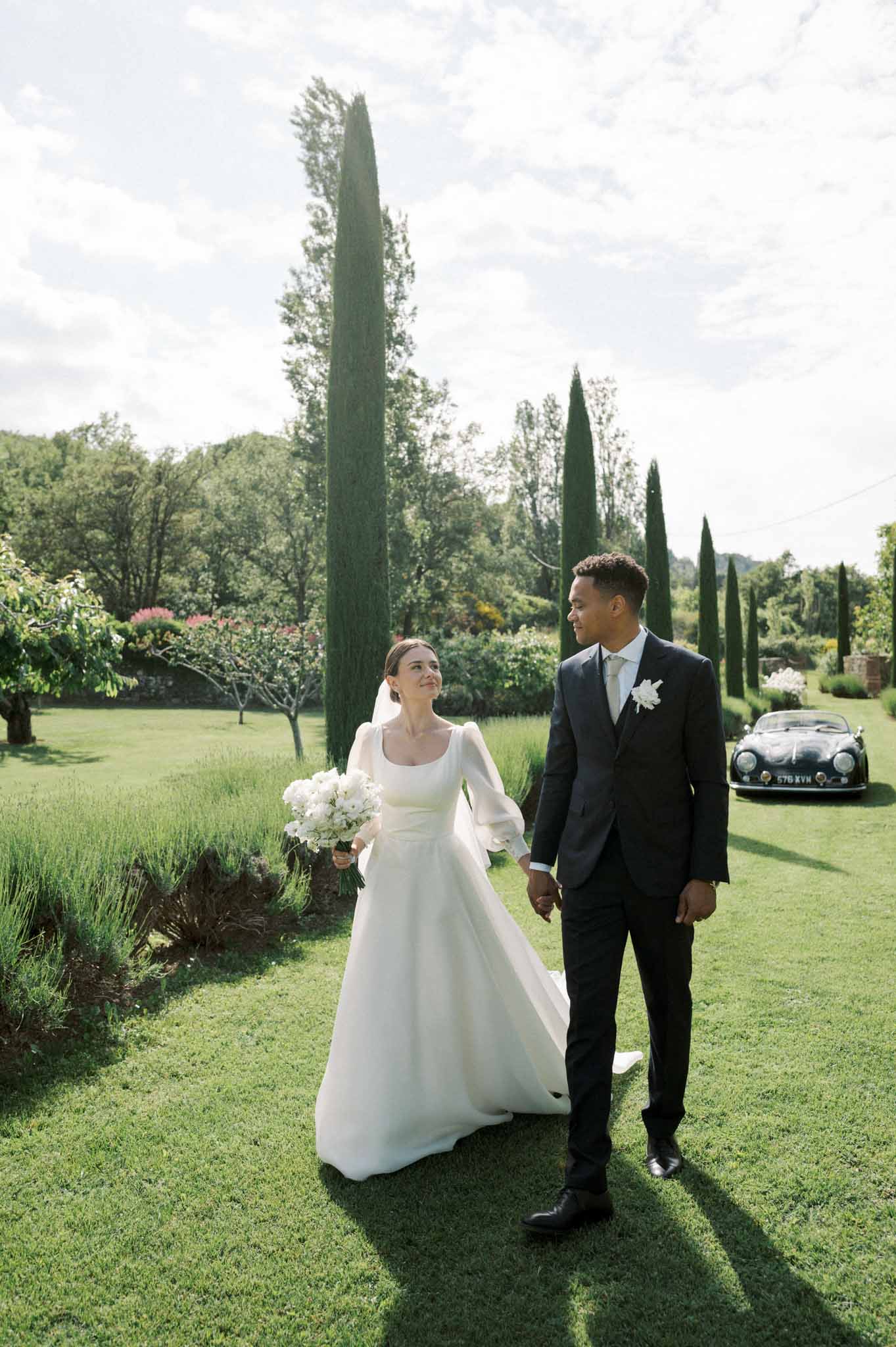 Couple walking on lawn with vintage Porsche and cypress trees, bride in puff-sleeve gown