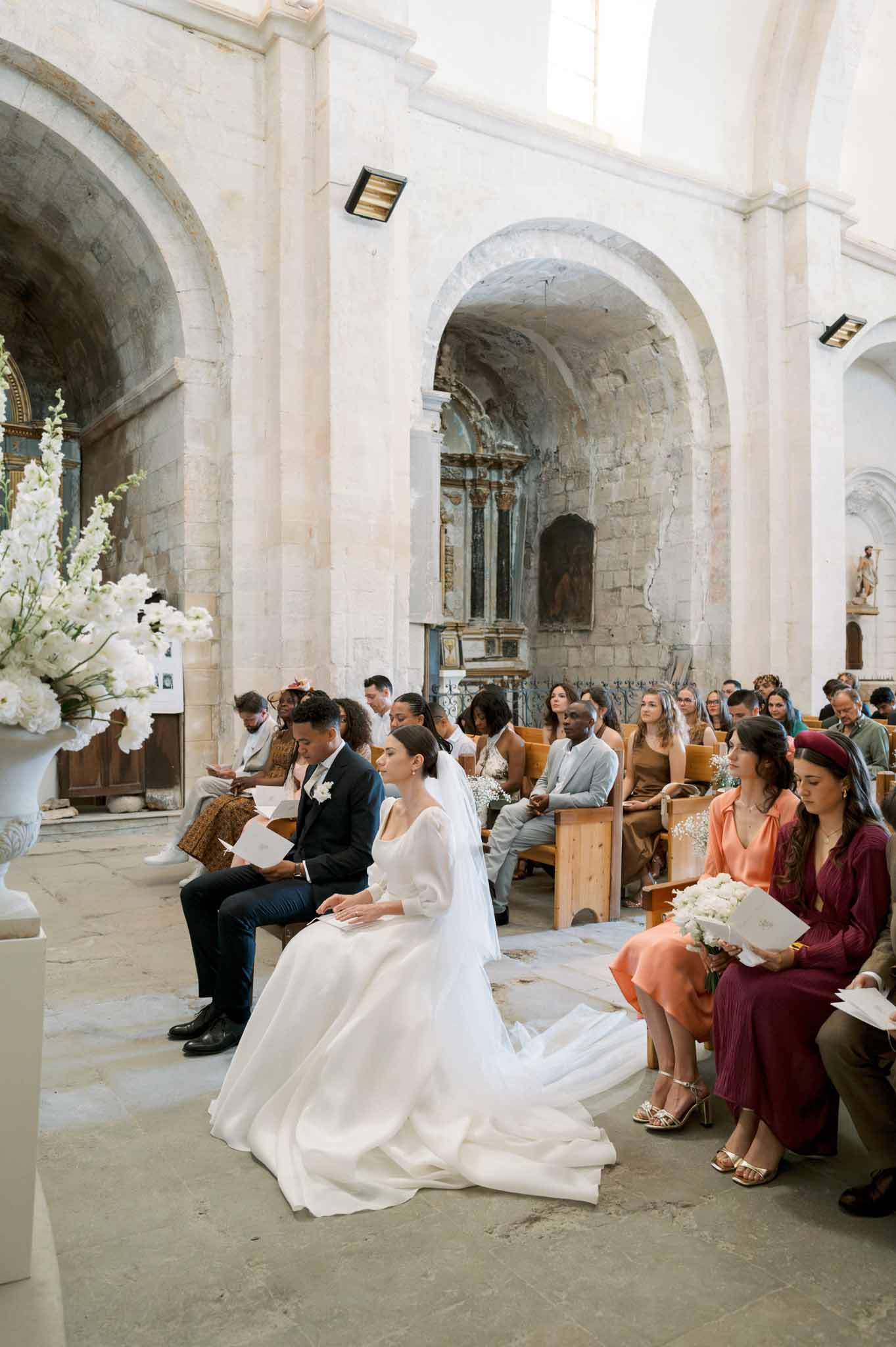 Bride and groom seated inside stone chapel with bridesmaids in terracotta and burgundy dresses and white floral arrangements