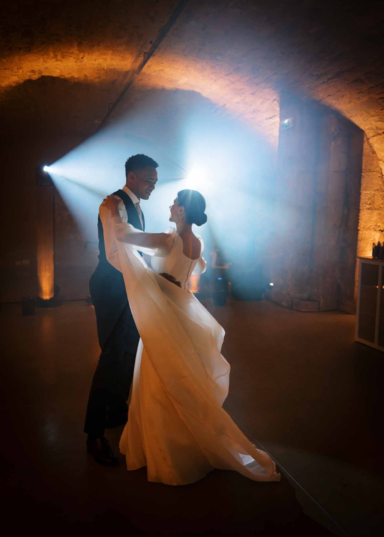 Bride and groom sharing first dance in a vaulted stone cellar with dramatic blue and amber lighting