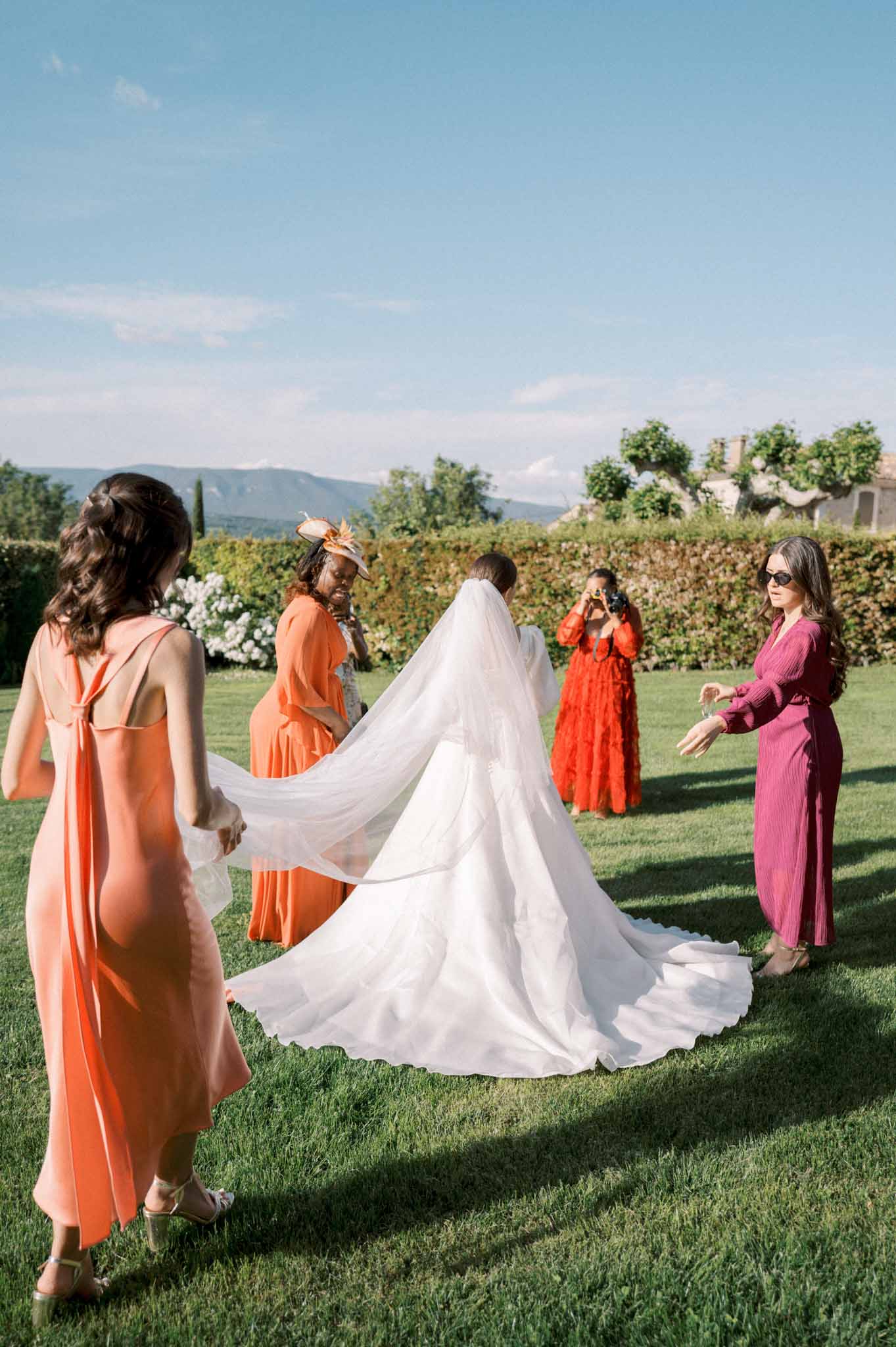 Four women in jewel-toned dresses arrange bride's cathedral veil and train on garden lawn before ceremony