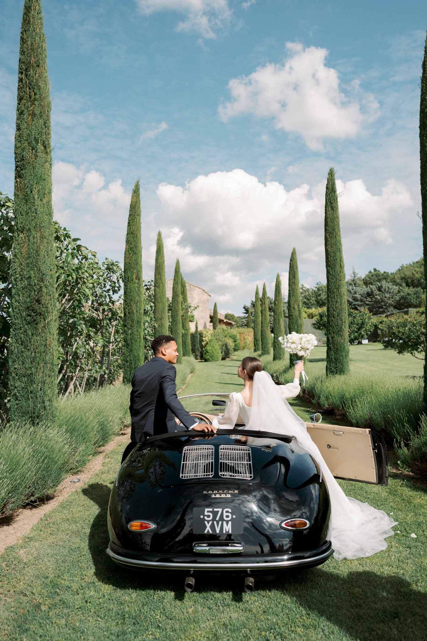 Couple in black Porsche 356 convertible on cypress-lined drive bride raising white bouquet cathedral veil trailing