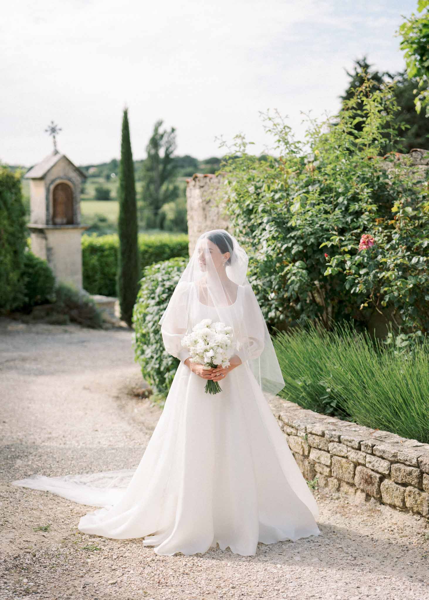 Bride in white ball gown with puff sleeves and cathedral veil holding white bouquet on gravel path near stone chapel