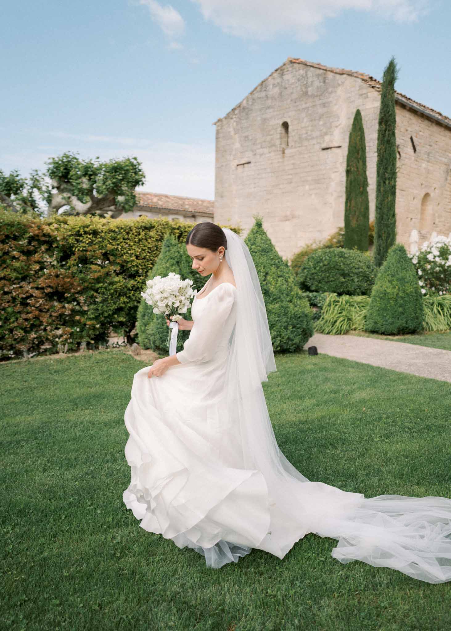 Bride in long-sleeve white gown with cathedral train and white bouquet before limestone abbey and topiary garden