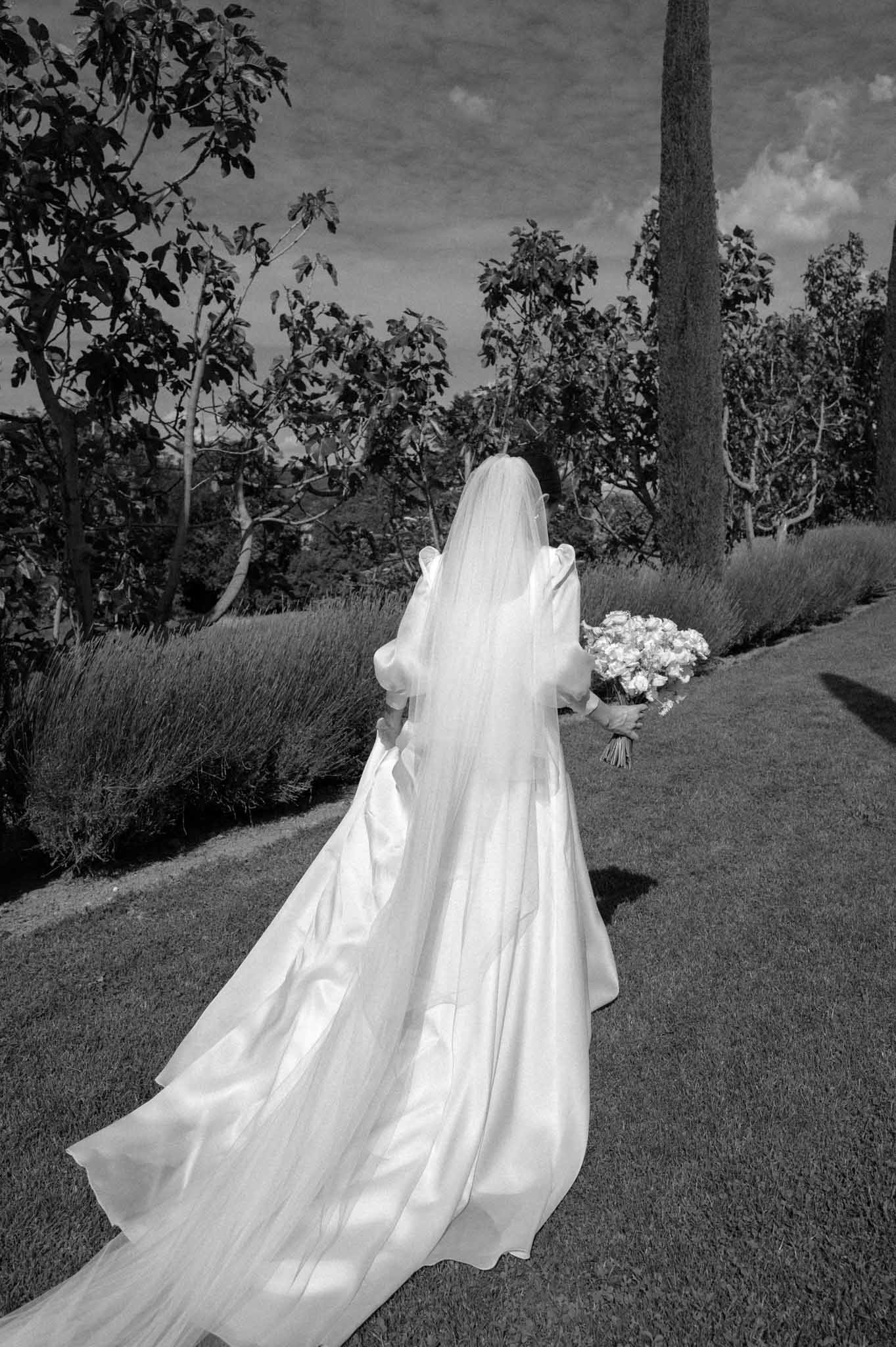 Black-and-white photo of bride walking away across a Provencal garden in puff-sleeve gown with cathedral train