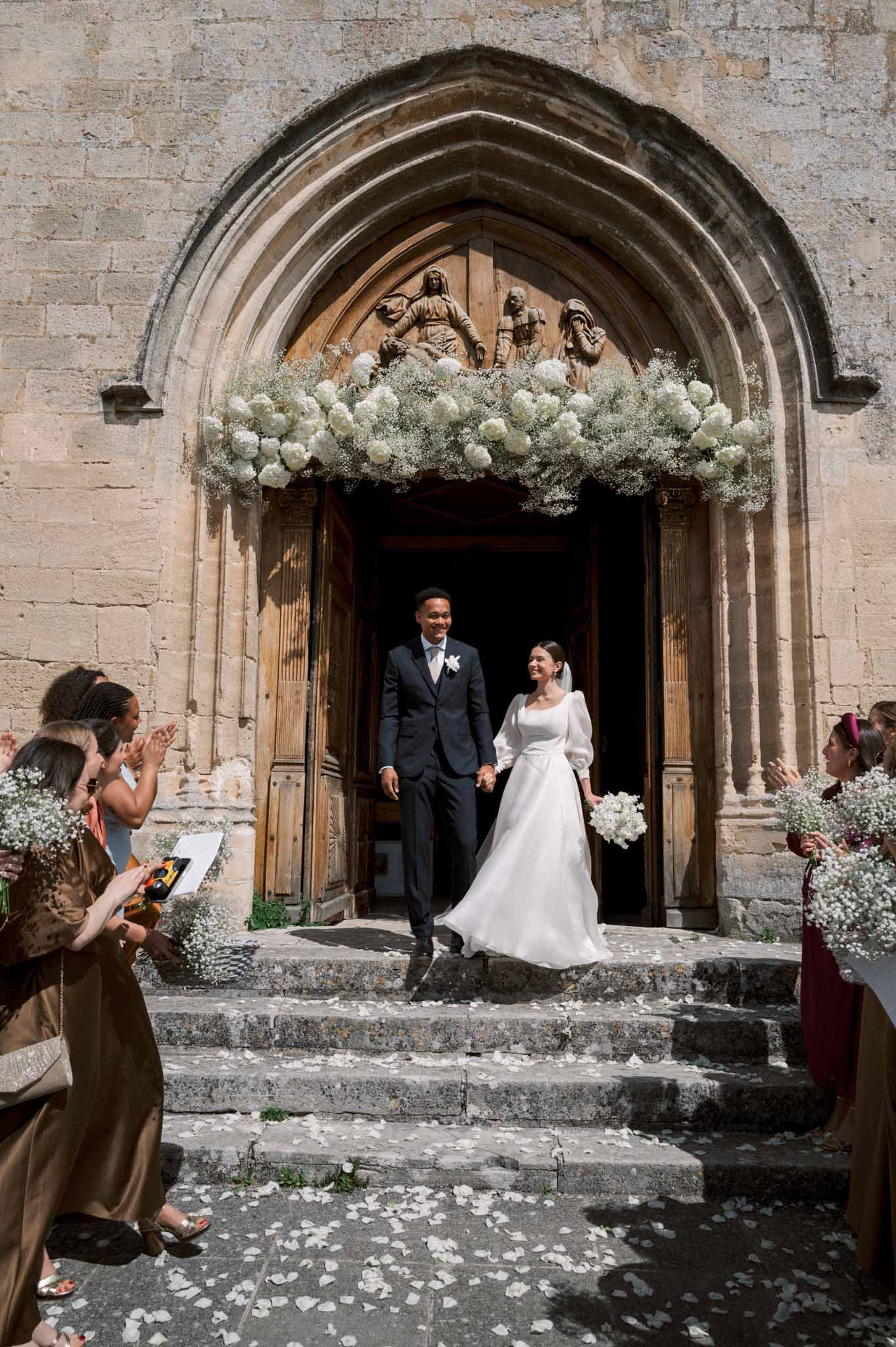 Bride and groom exiting stone church under white hydrangea arch as guests applaud