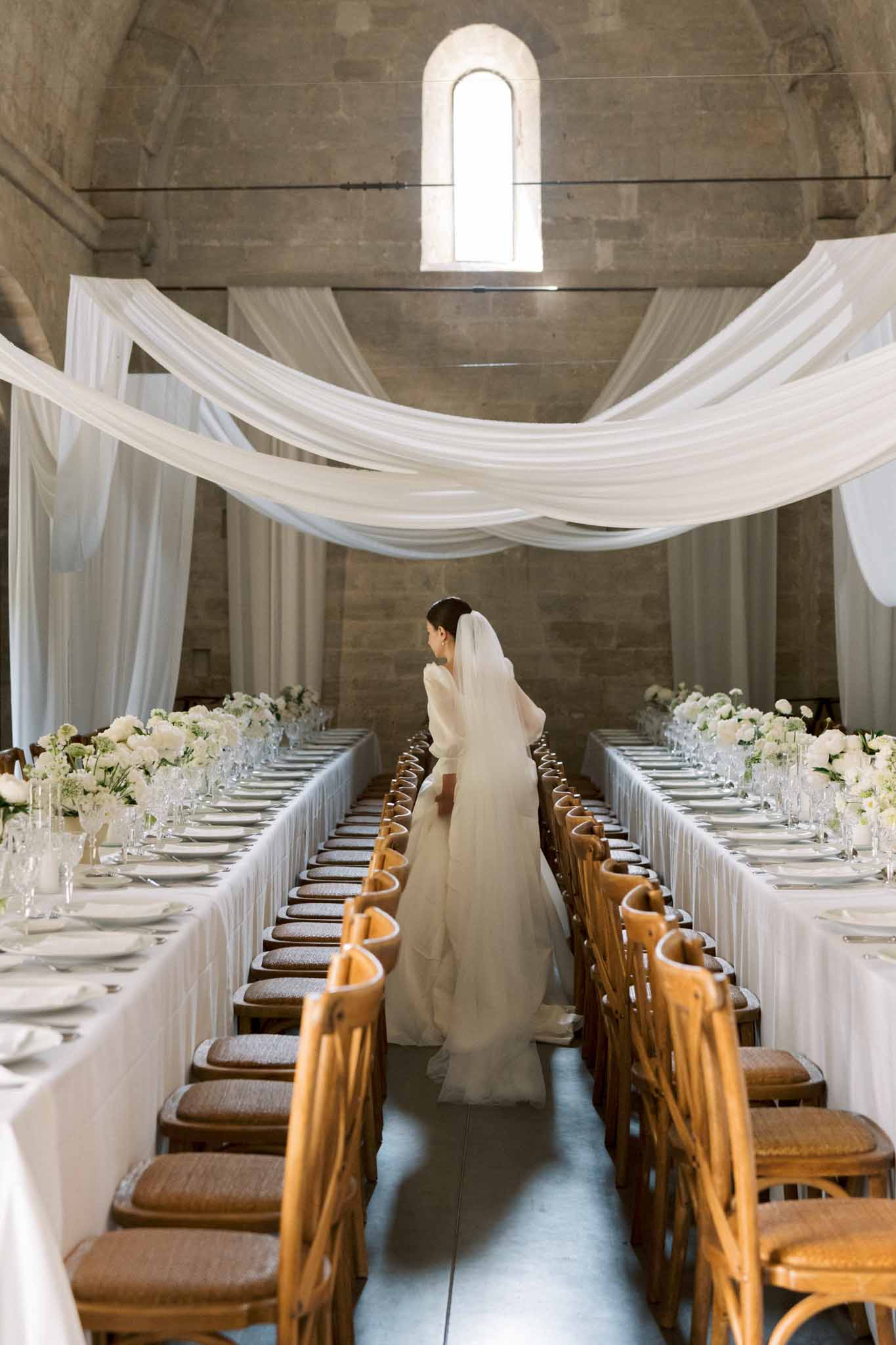 Bride with cathedral veil between two banquet tables with white roses in draped stone-vaulted abbey hall