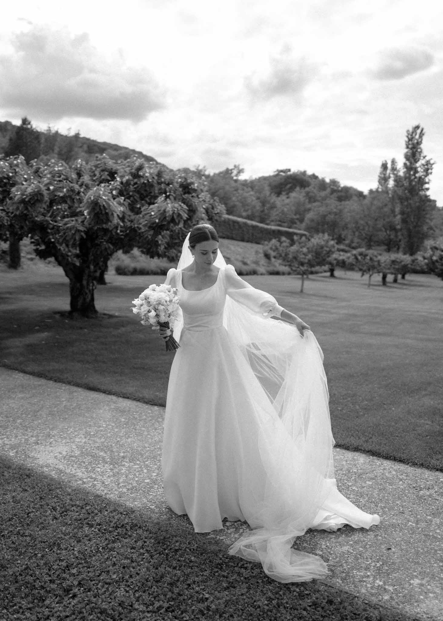 Black and white full-length bridal portrait with bride lifting cathedral veil and holding bouquet on garden path