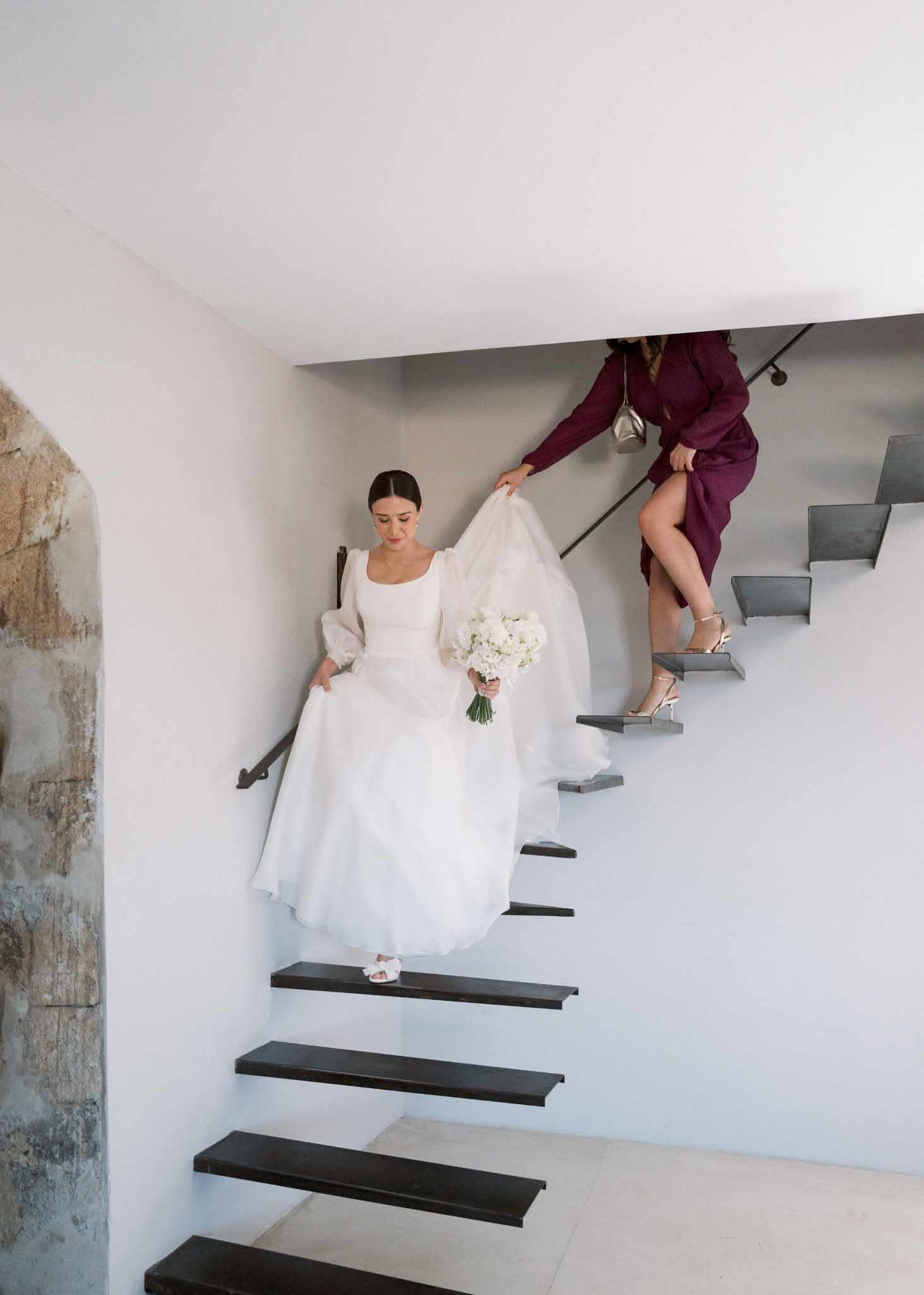 Bride descending floating staircase with white hydrangea bouquet and companion in burgundy dress