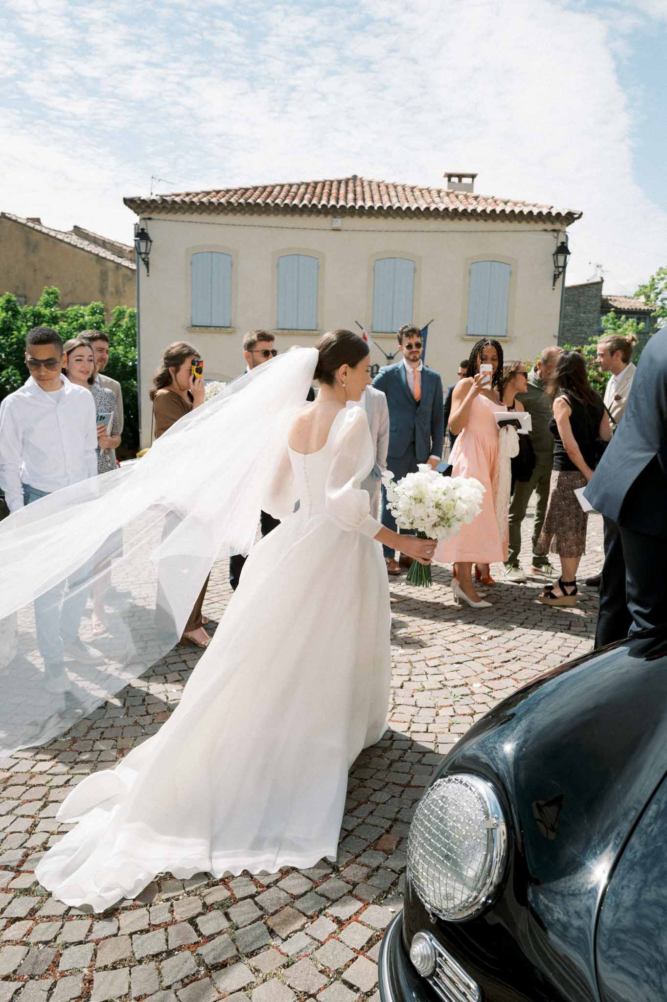 Bride in white long-sleeve gown and cathedral veil walks through cobblestone village square as guests photograph her