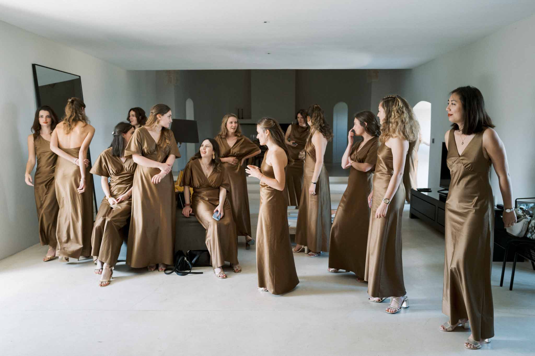 Eight bridesmaids in bronze-gold satin dresses gathered in modern white-walled room with floor mirror