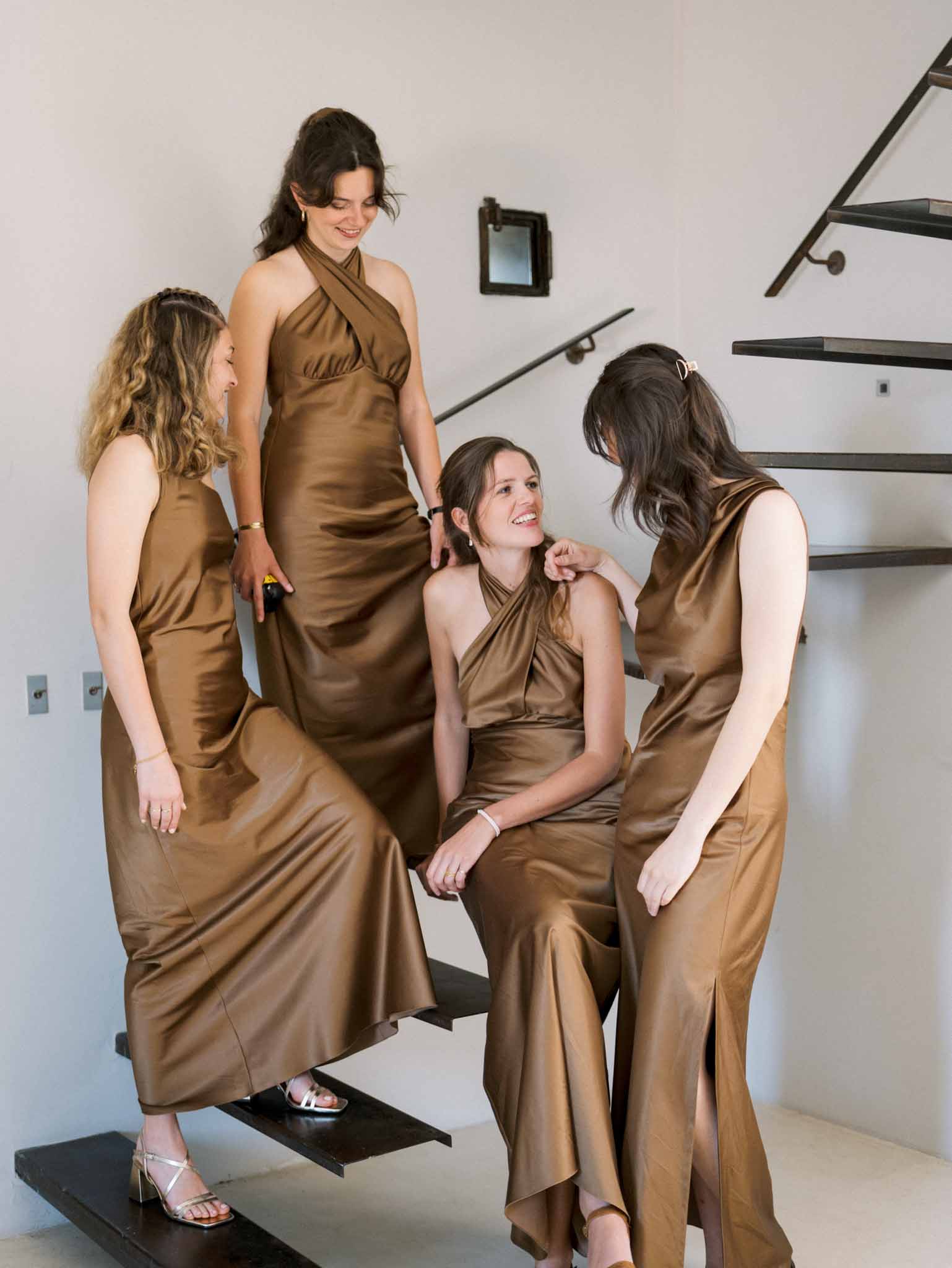 Four bridesmaids in bronze-brown satin floor-length dresses laughing on modern metal-and-glass staircase