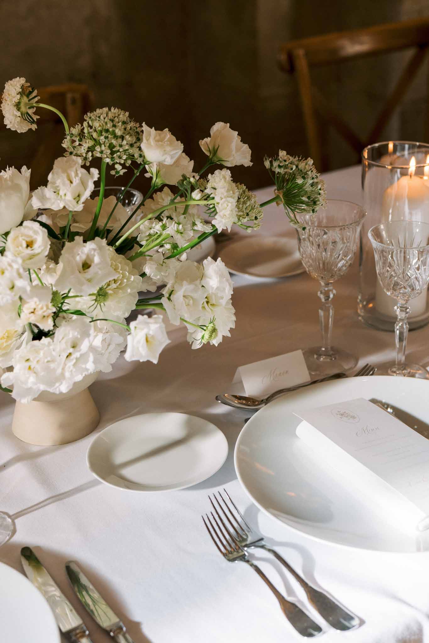 Close-up of reception place setting with cream ceramic vase of white lisianthus, crystal glasses, and calligraphy place card