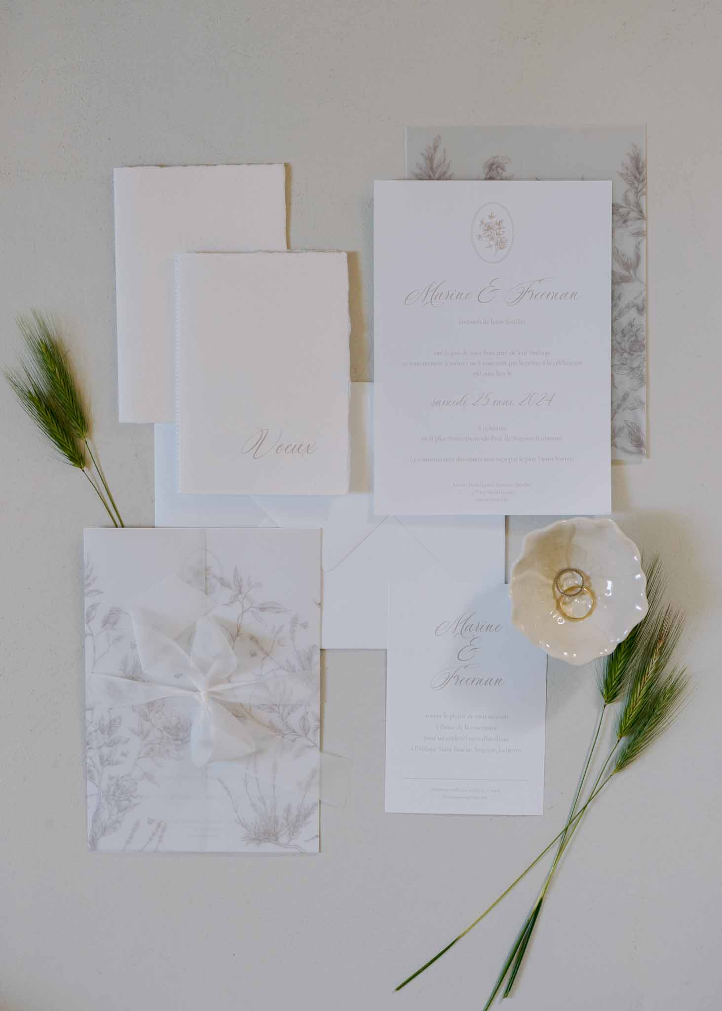French wedding stationery flat lay with gold calligraphy invitations, vellum overlay, gold rings, and wheat stems