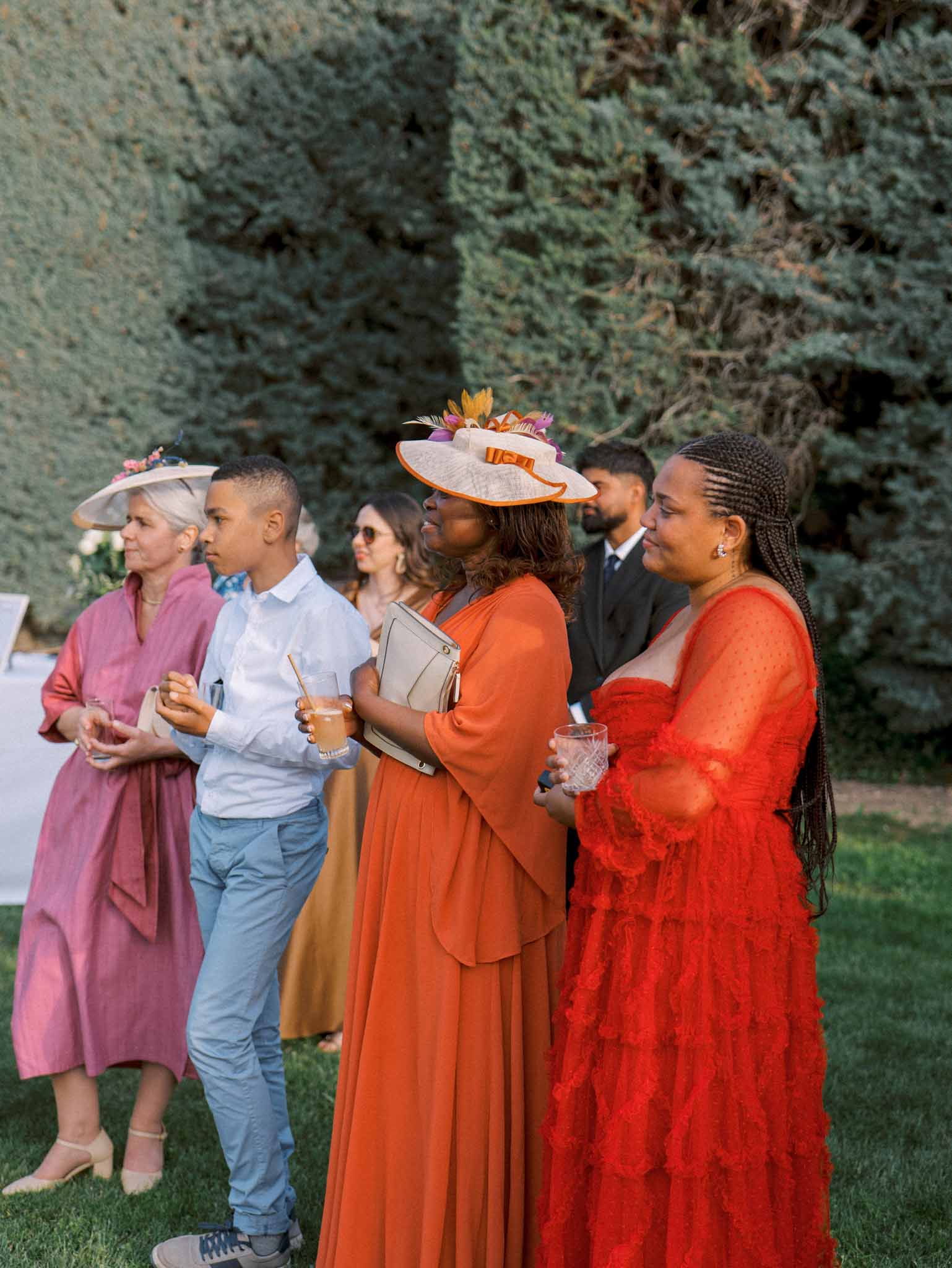 Wedding guests in rose, burnt orange, and red dresses with wide-brim hats watching from hedged garden