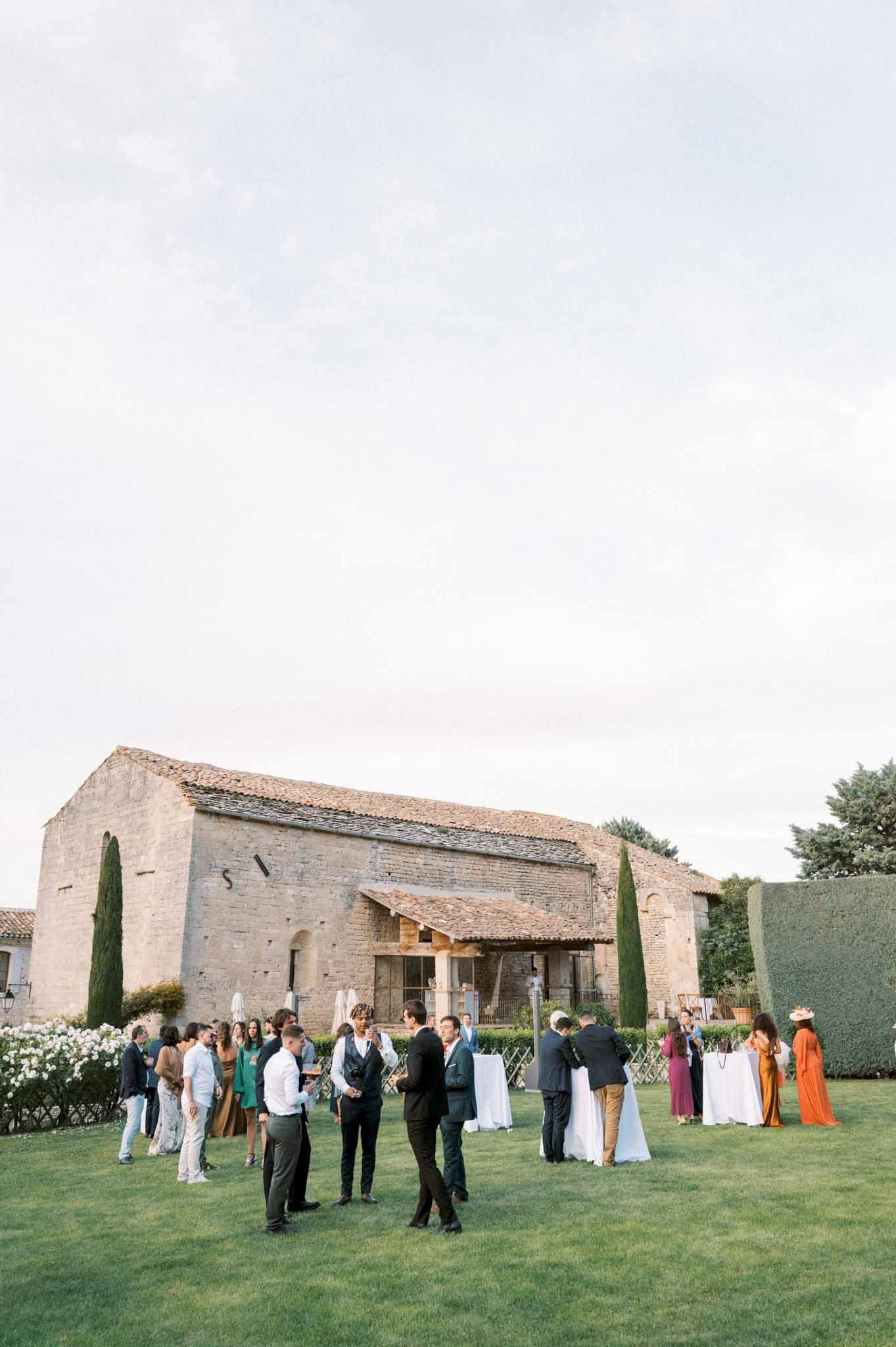 Guests mingling at cocktail tables on lawn before Provencal stone building with cypress trees