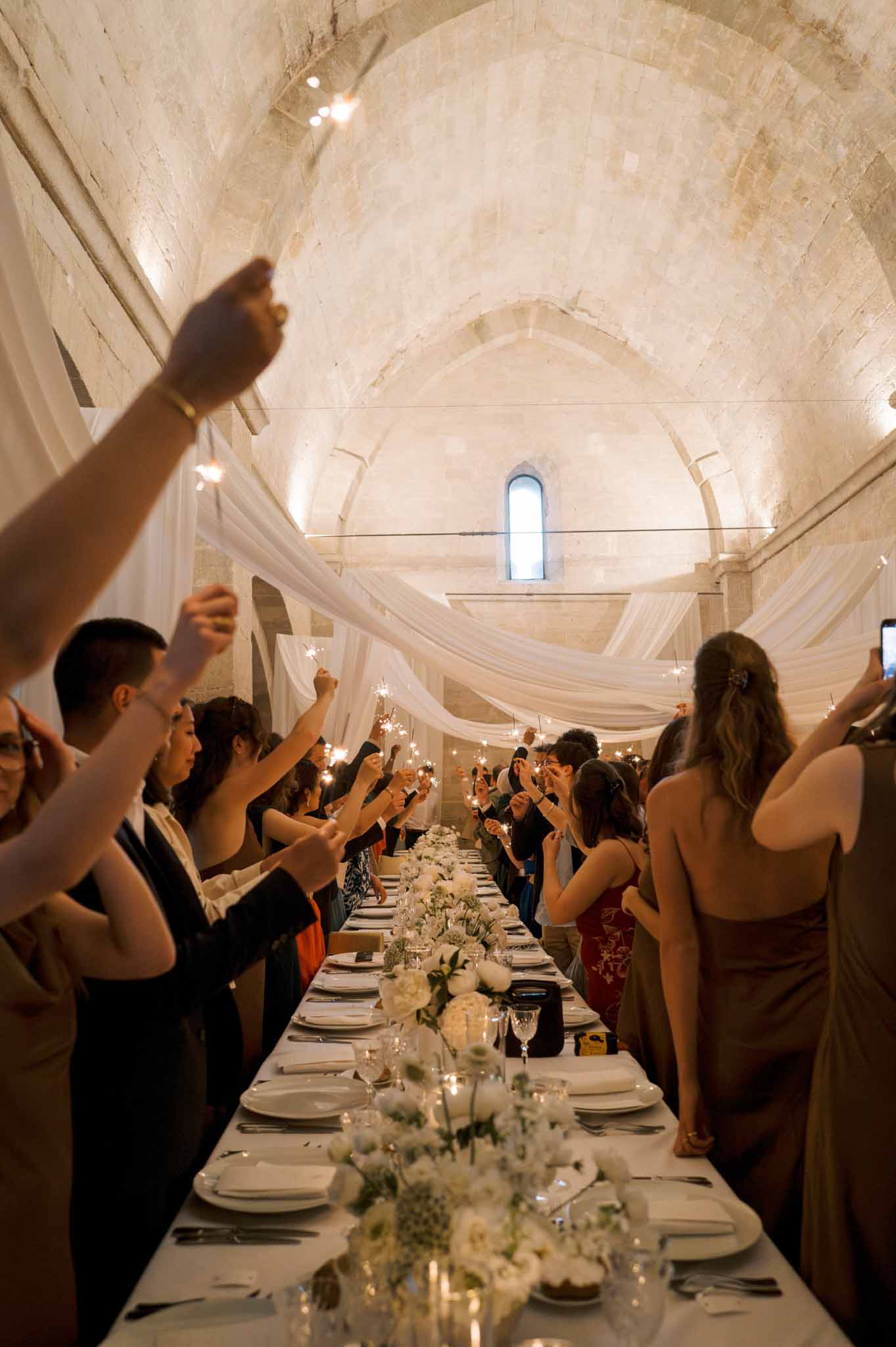 Guests hold sparklers over long banquet table inside medieval stone abbey with vaulted ceilings and white draping