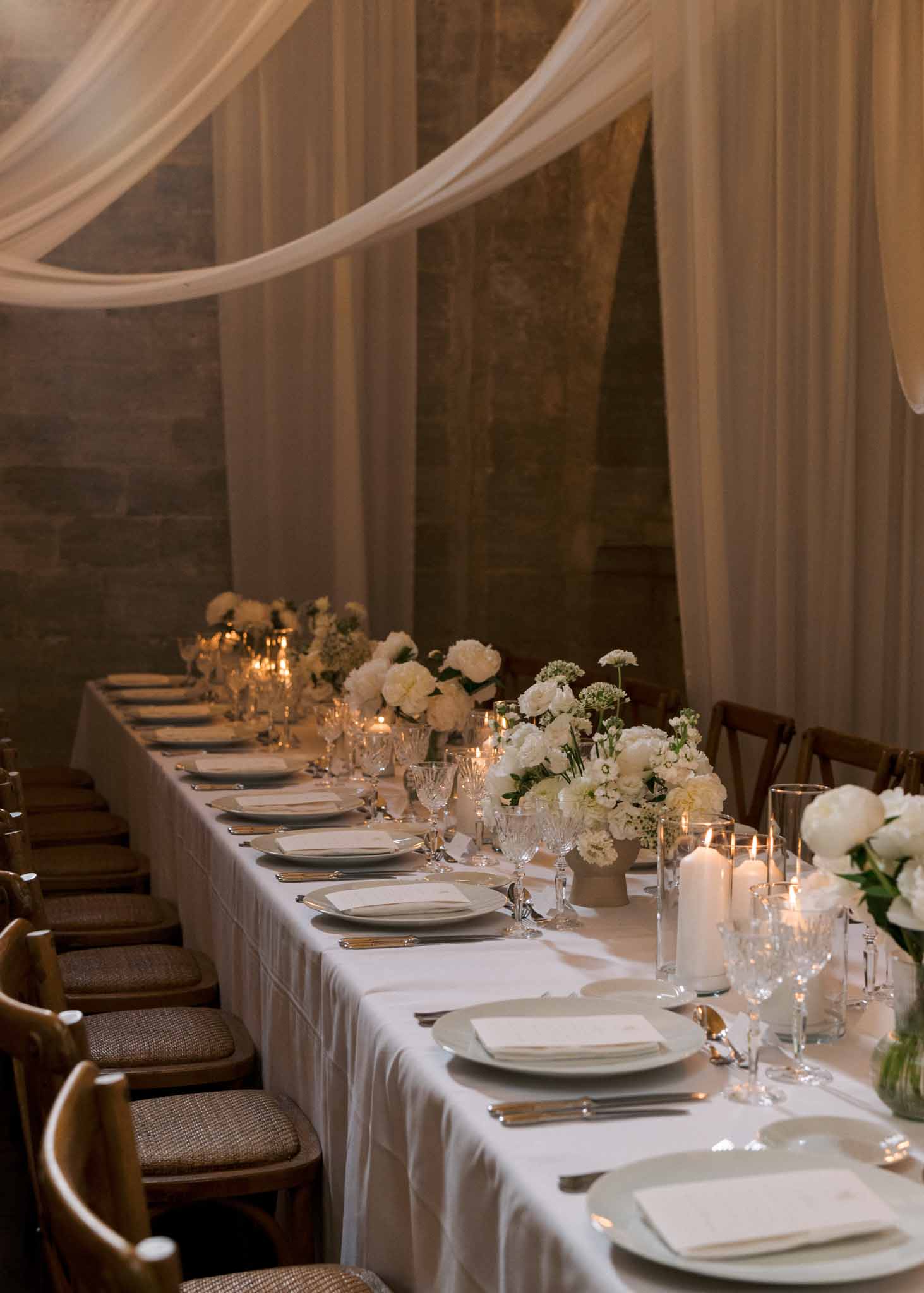 Indoor reception with white peonies, chiffon ceiling draping, and cross-back chairs on stone walls