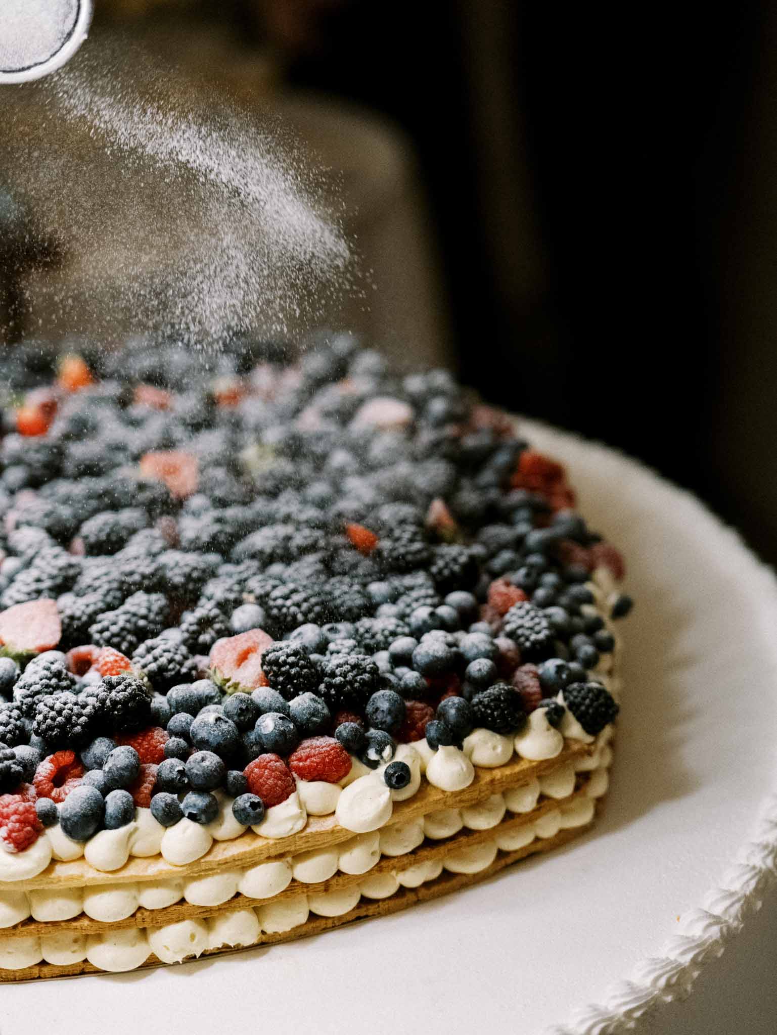 Berry tart cake being dusted with powdered sugar on white linen against dark background