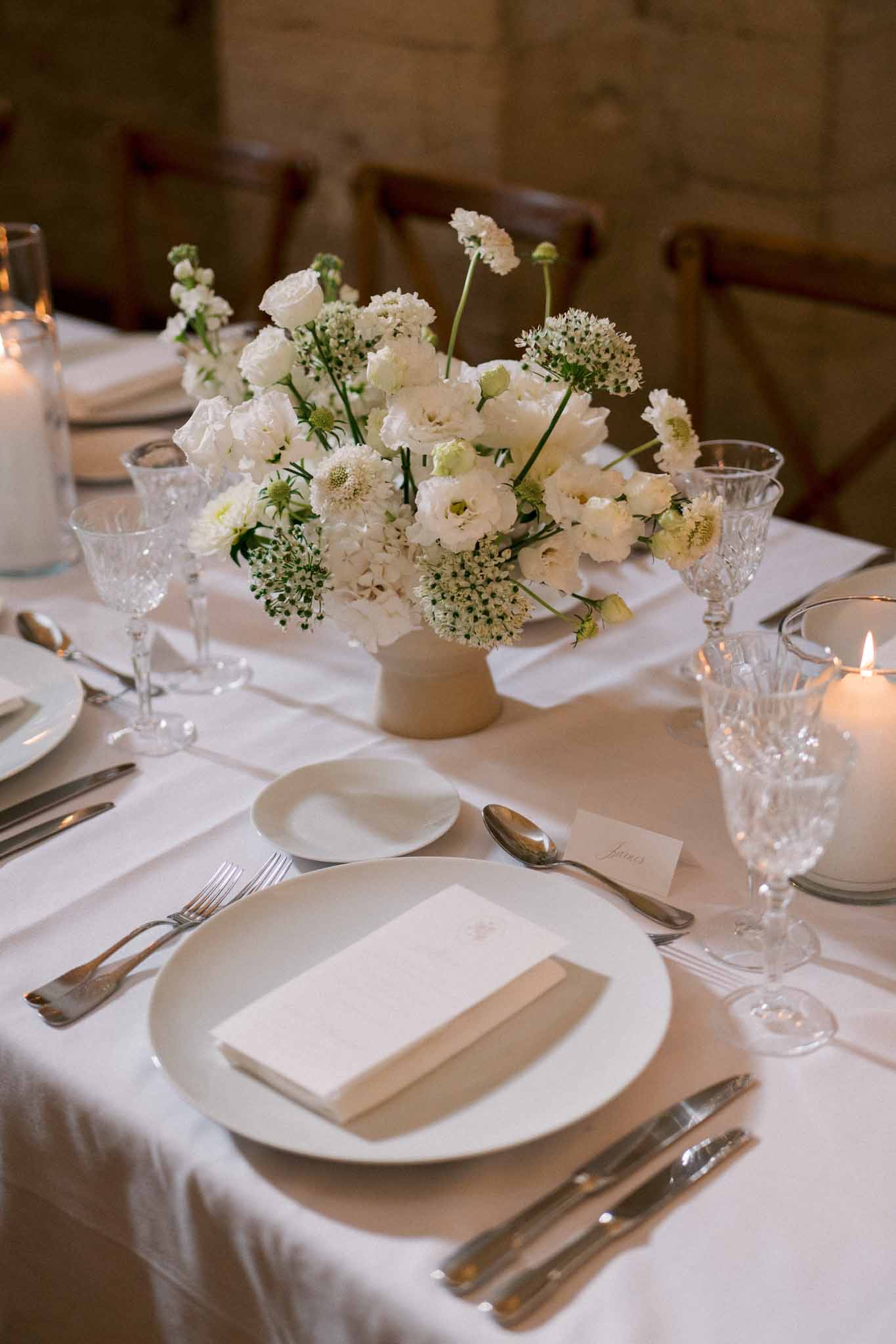 Close-up of reception table with all-white floral centerpiece in beige pedestal vessel, crystal goblets, and pillar candles