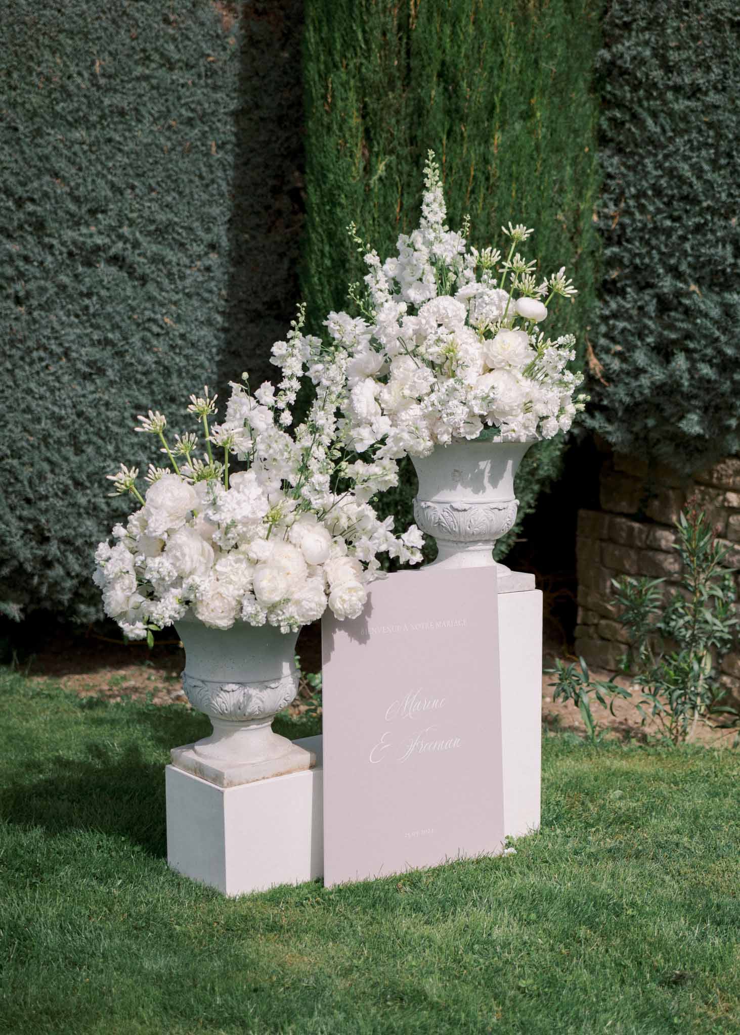 Two stone urns with white peony and delphinium arrangements flanking blush pink calligraphy welcome sign on lawn