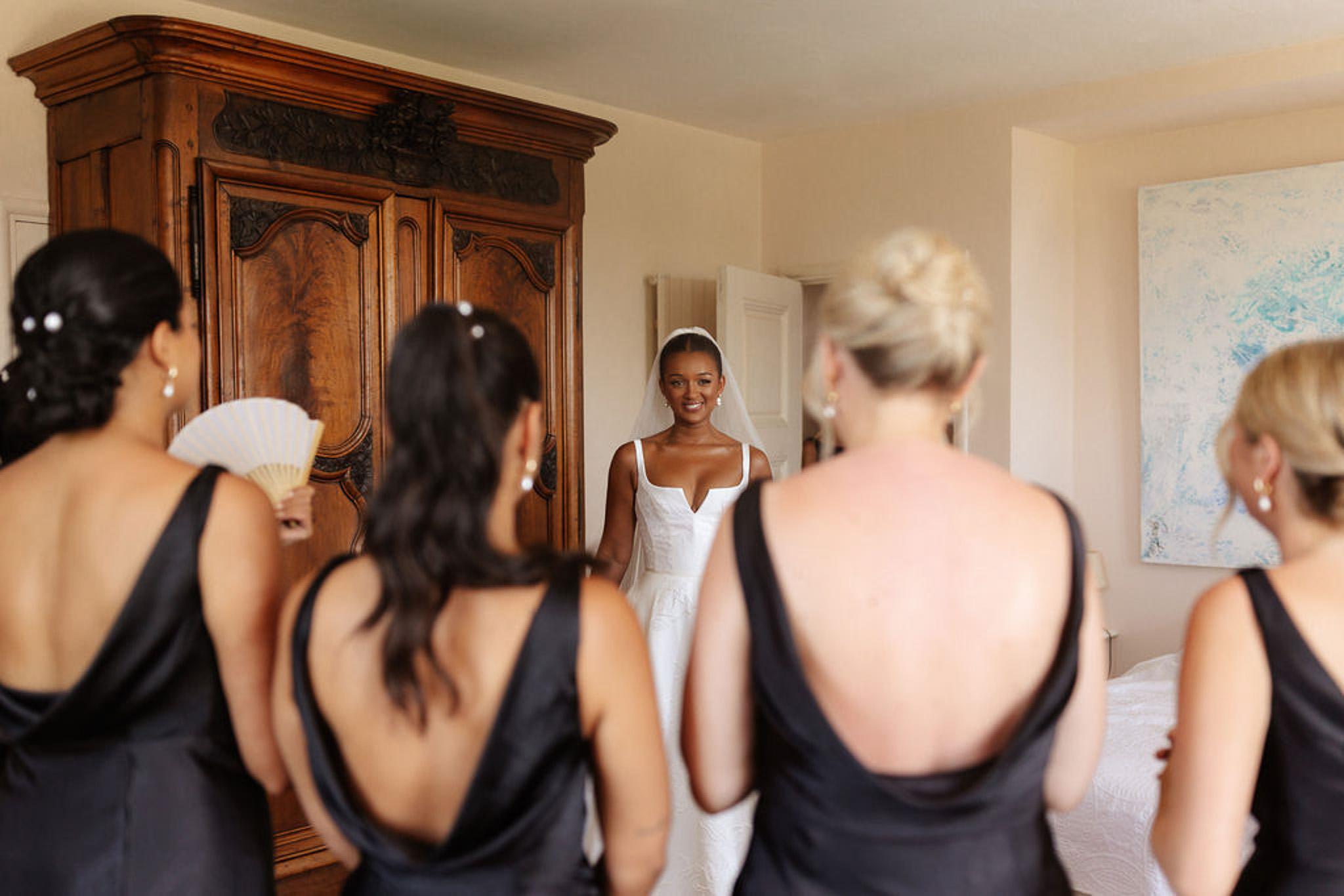 Bride in ivory dress with bridesmaids in black dresses getting ready in bedroom with wooden armoire