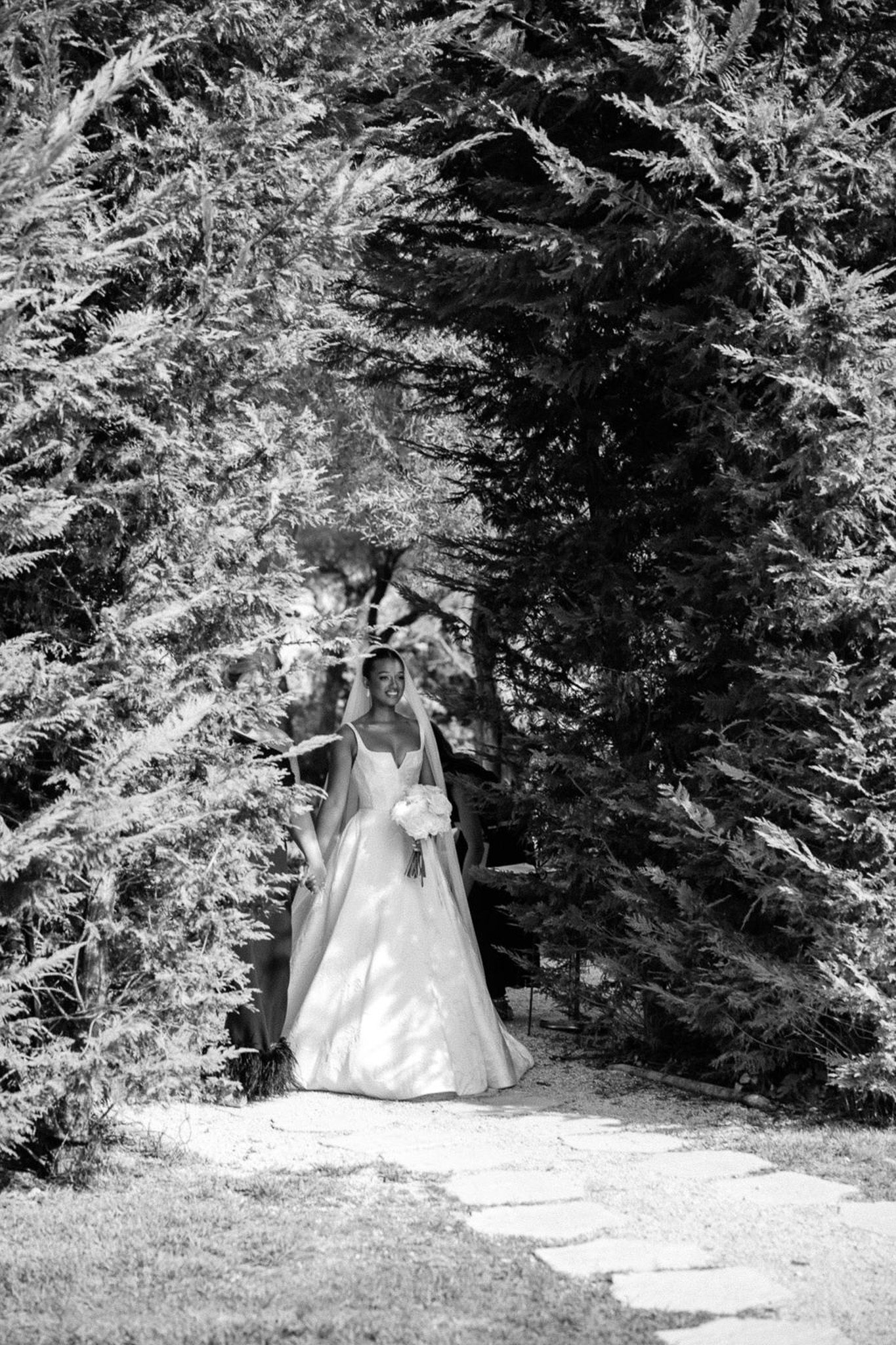 Bride in ivory wedding dress standing on stone pathway framed by evergreen trees in black and white portrait